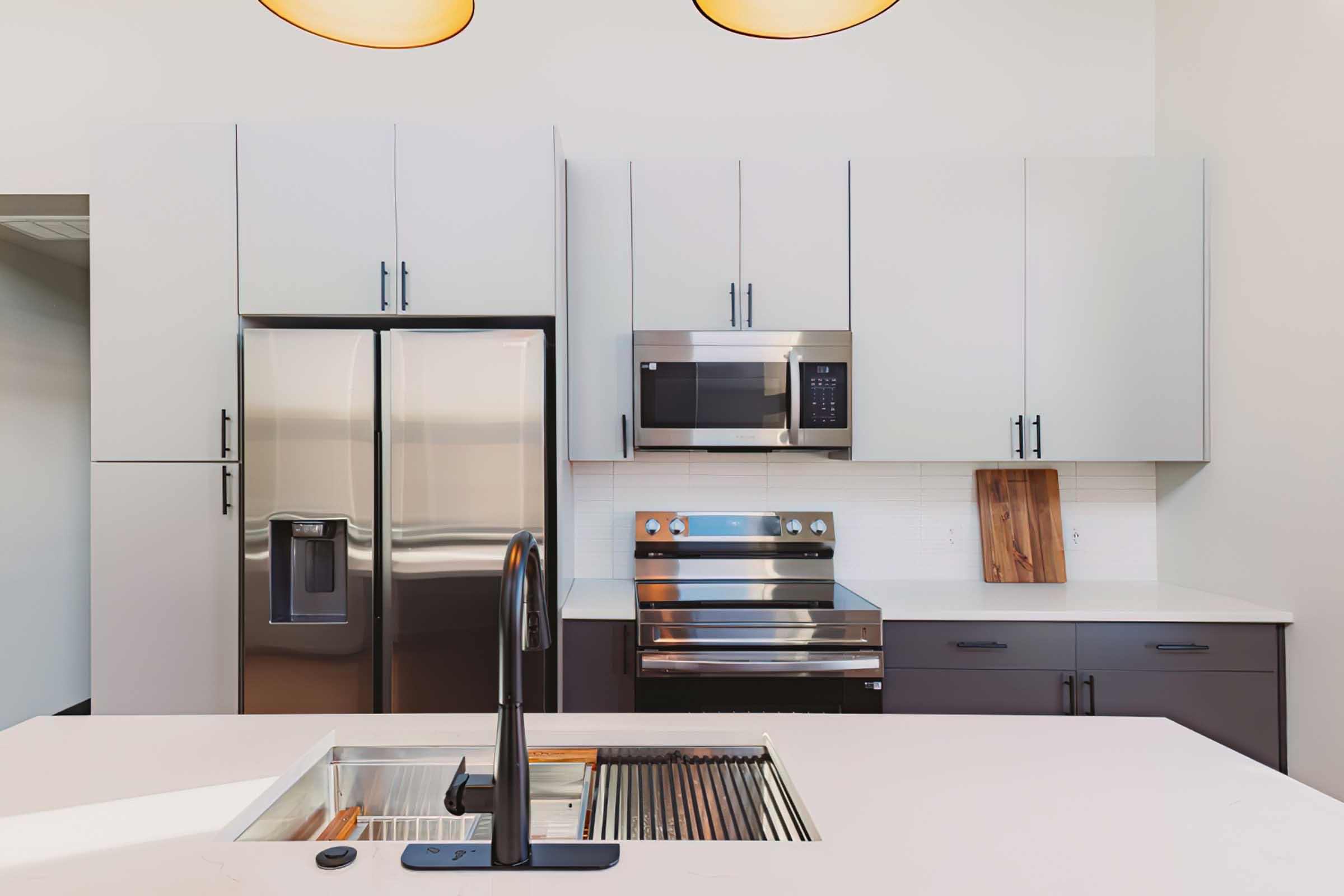 Modern kitchen featuring stainless steel appliances, including a refrigerator, microwave, and oven. The kitchen has light gray cabinets, a spacious white countertop with a built-in sink, and a wooden cutting board. Soft lighting from overhead pendant lamps enhances the contemporary design.
