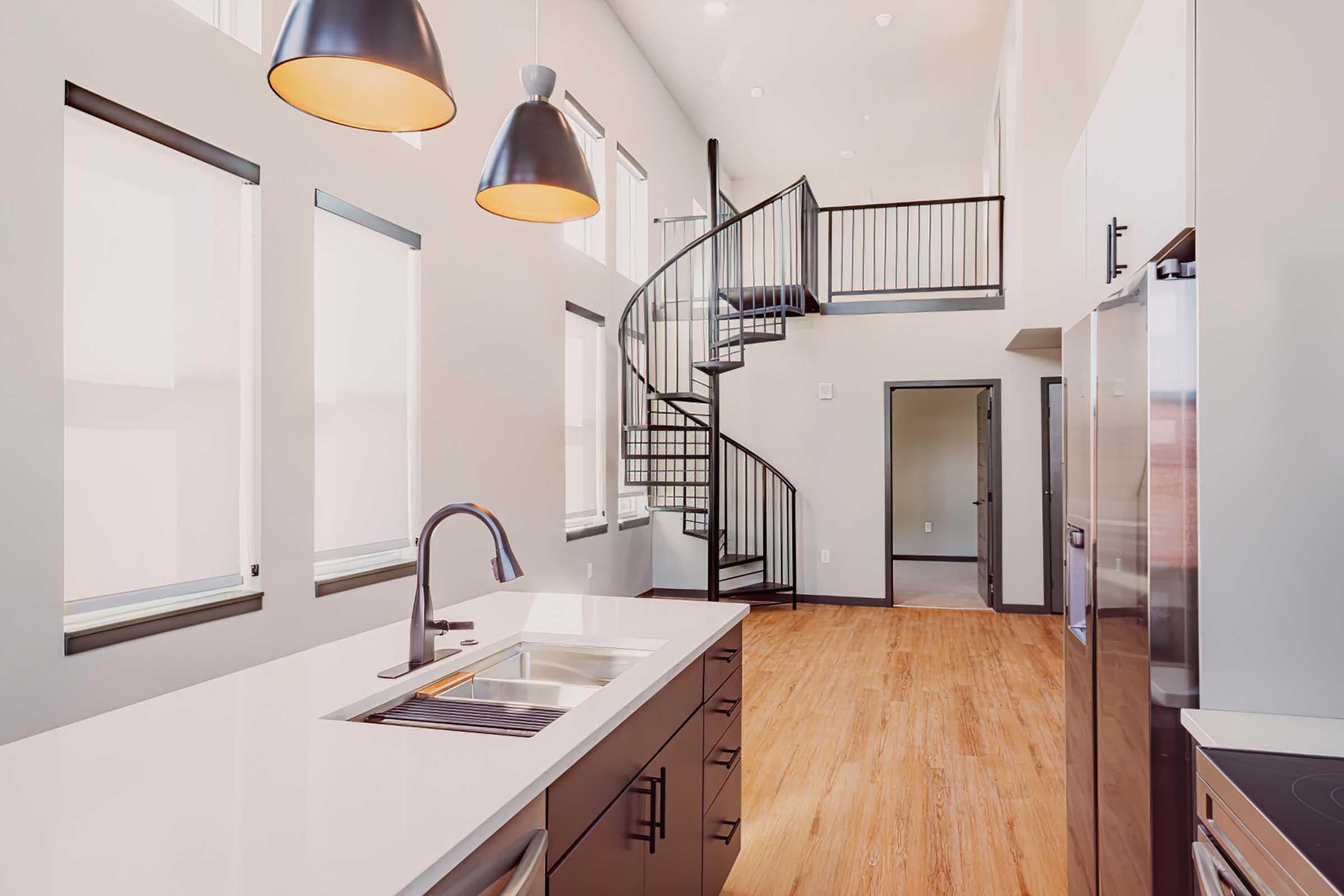 Modern kitchen with a sleek design featuring a large island and stainless steel appliances. There are tall windows allowing natural light, and a spiral staircase leads to an upper level. The floor is hardwood, complementing the neutral color scheme of the space.