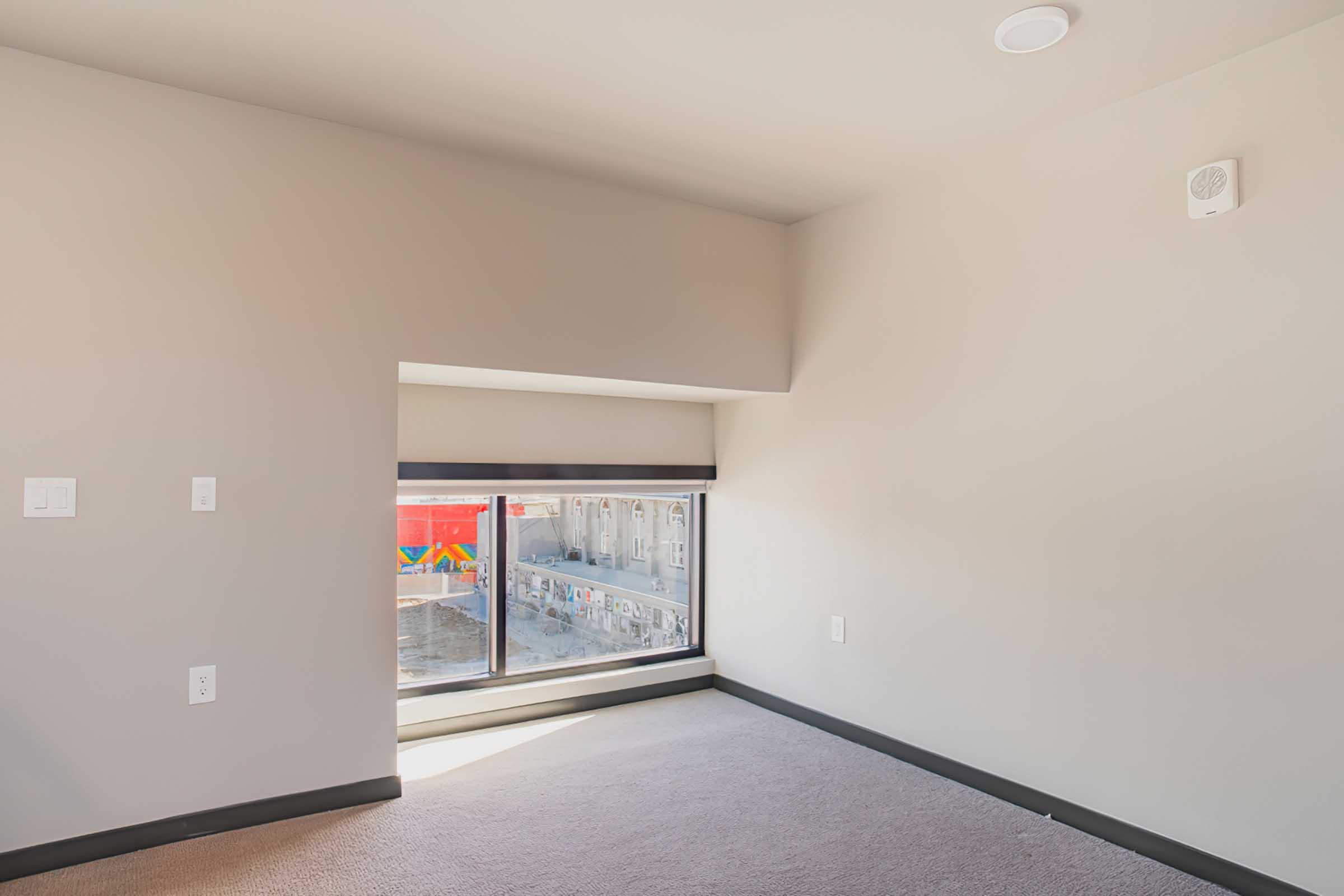 A minimalist room featuring light gray walls and carpet, with a large window letting in natural light. The window has a view of a colorful outdoor mural. There are two electrical outlets on the wall, and a small vent is visible above the window. The overall aesthetic is modern and uncluttered.