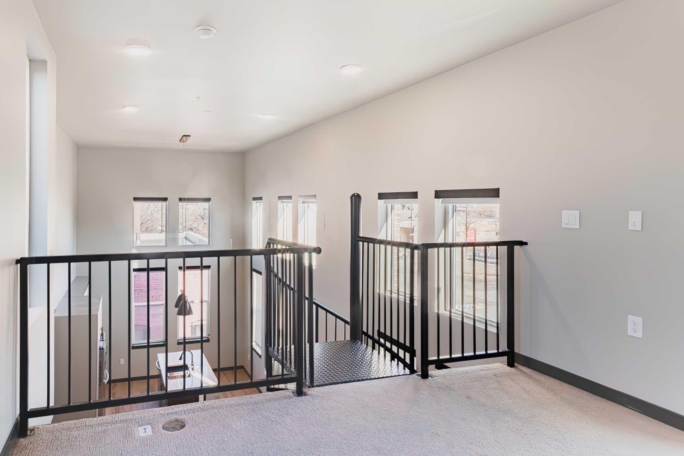 Interior view of a modern staircase landing with a black metal railing. The walls are light gray, and there are several windows allowing natural light to enter. The space is empty, featuring carpeted flooring and minimalist lighting fixtures.