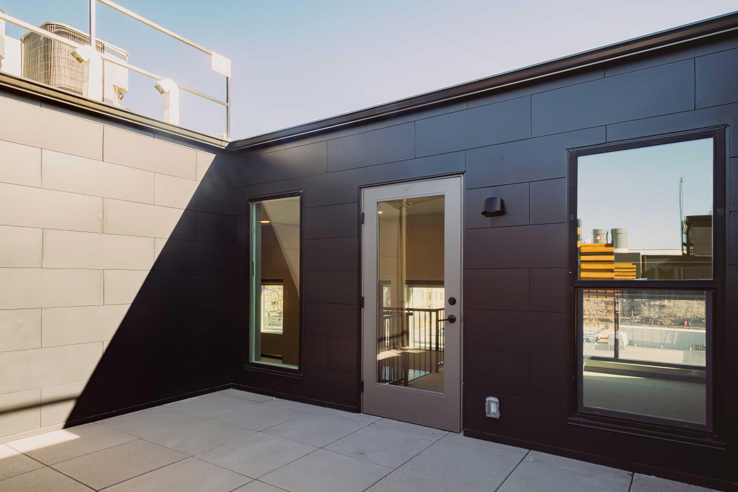 A modern rooftop terrace featuring black exterior walls, large windows, and a door leading to an interior space. The area has a spacious layout with a light-colored floor and a view of surroundings. Bright sunlight creates shadows, highlighting the architectural design.