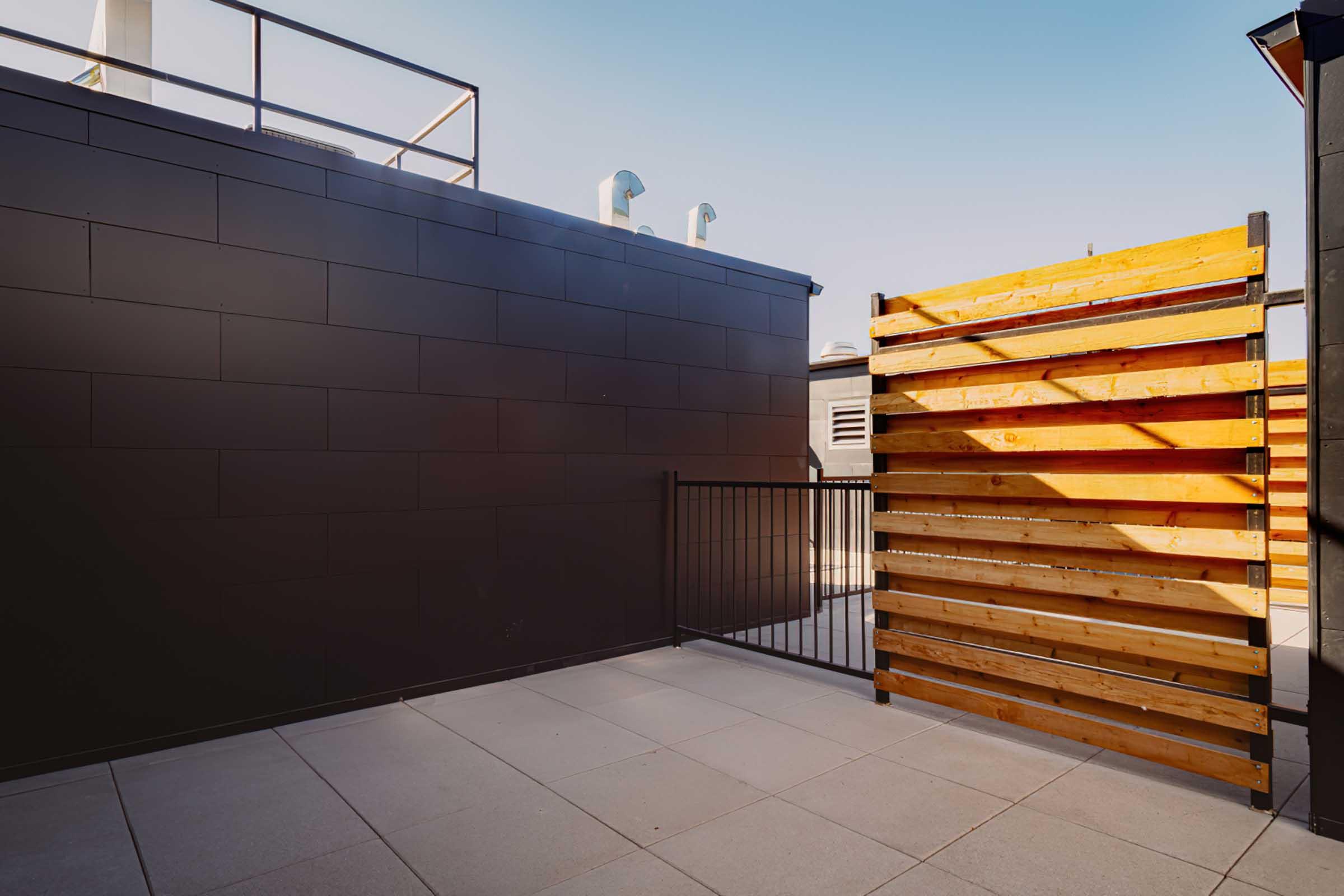 A rooftop space featuring a modern design with a black wall on one side, a wooden slatted fence on another, and light-colored paving stones. The area is open and bright, with clear skies above, suitable for outdoor relaxation or gatherings.