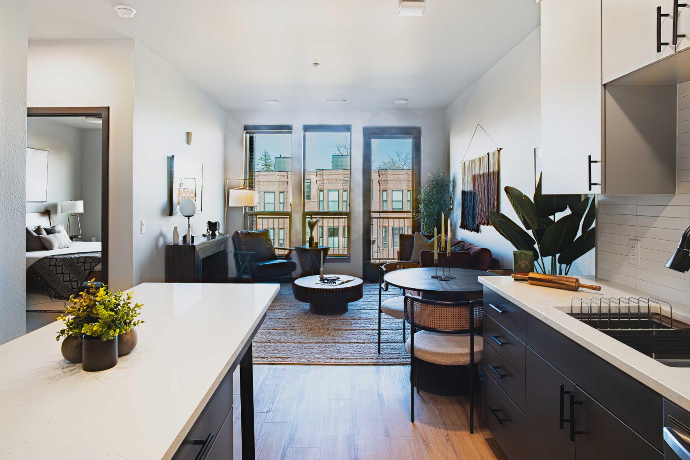 A modern, stylish apartment interior featuring an open kitchen and living area. The kitchen has dark cabinets and a marble countertop. The living space includes a round coffee table, a comfortable chair, and large windows with natural light. Decorative plants and wall art complete the cozy atmosphere.