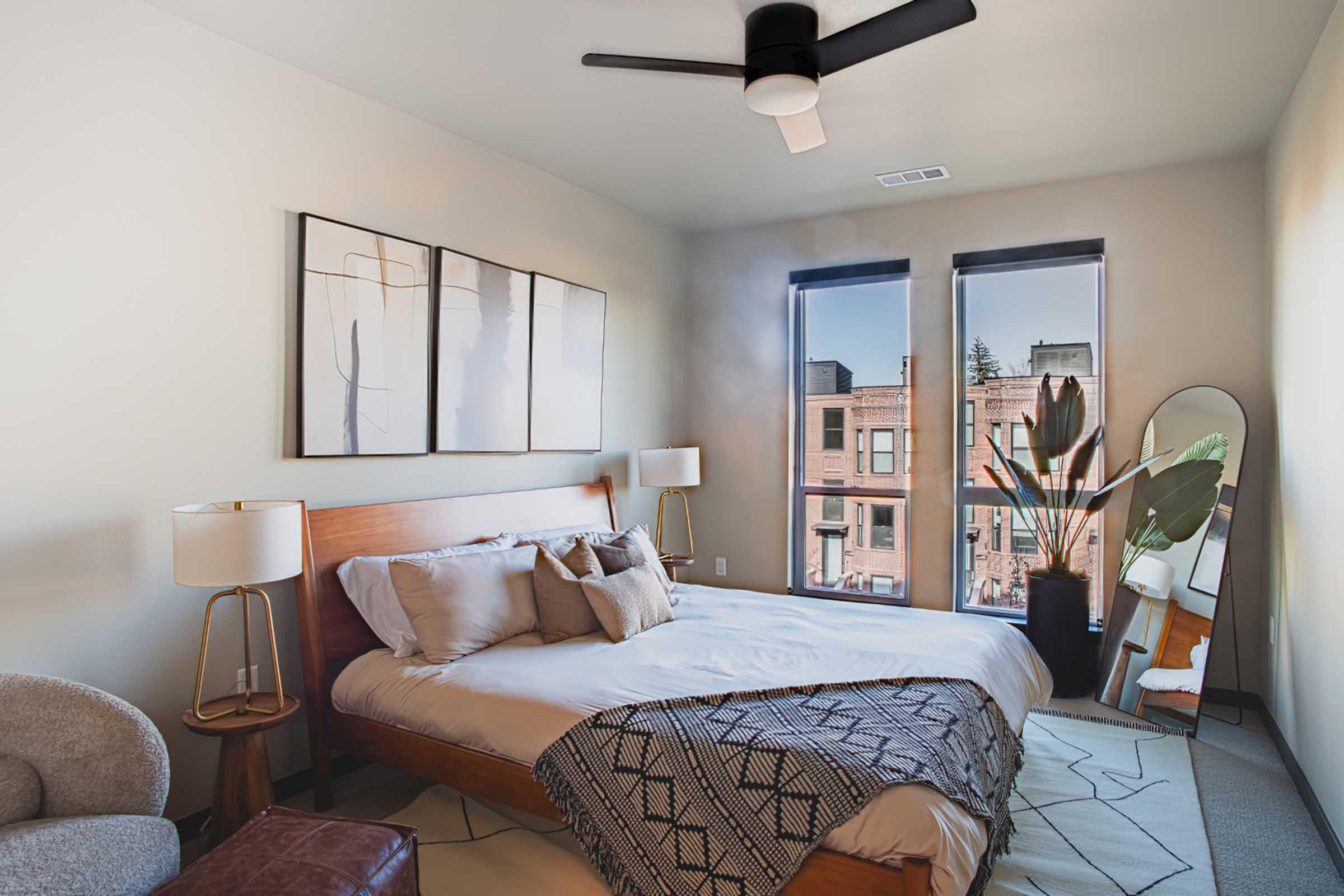 Modern bedroom featuring a wooden bed with decorative pillows and a cozy blanket. Large windows allow natural light, showcasing a view of city buildings. A potted plant adds a touch of greenery, while stylish lamps and an accent chair complete the inviting atmosphere.