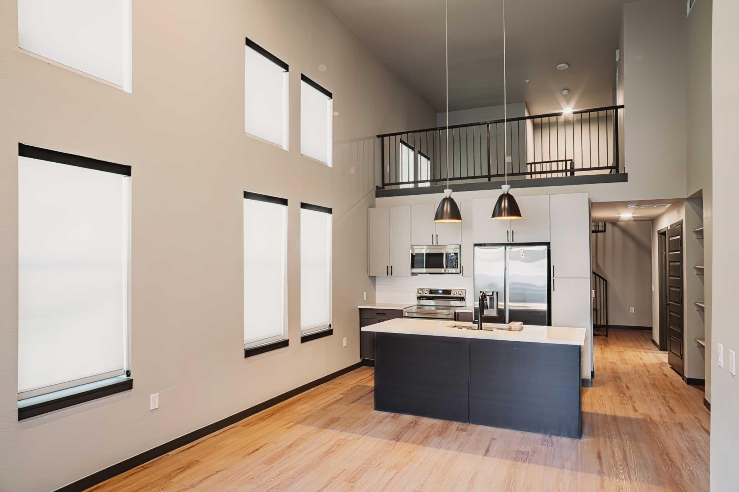 Modern kitchen and living area with high ceilings and large windows, featuring a central island with seating. Stainless steel appliances are in the kitchen, complemented by dark cabinetry. The space is bright with neutral wall colors and wooden flooring, leading to a staircase in the background.
