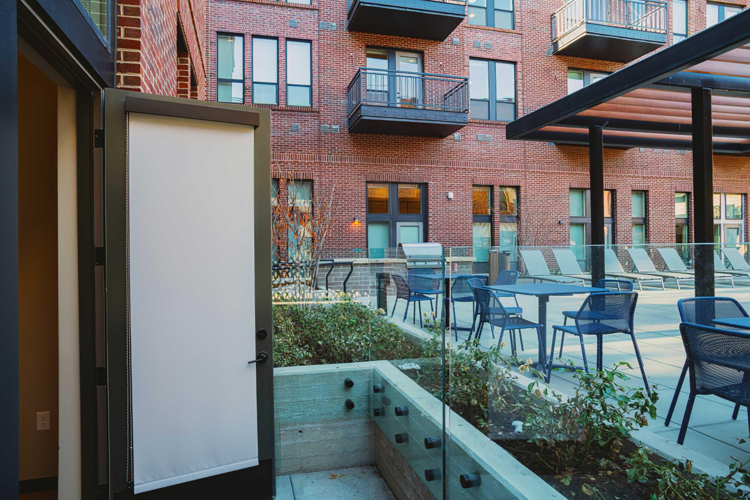 View of a modern apartment complex courtyard featuring a pool area with lounge chairs, surrounded by brick buildings. There is a glass railing and landscaping with plants visible in the foreground, along with a door leading into the apartment. Bright and inviting atmosphere.