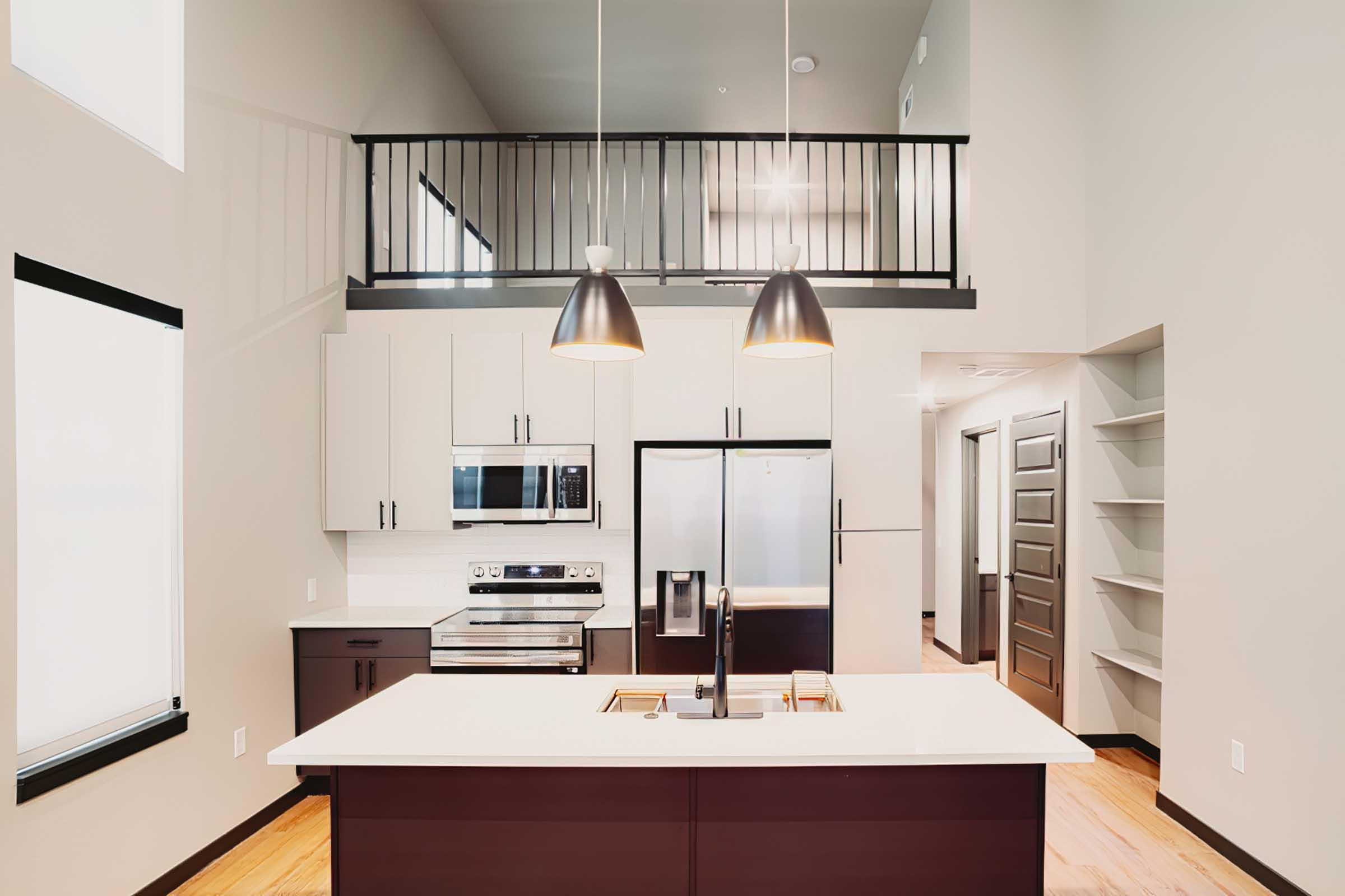 Modern kitchen with an open layout featuring a large island, stainless steel appliances, and dark cabinetry. The space includes overhead pendant lights and a loft area visible above. Natural light filters in through large windows, complementing the contemporary design and hardwood flooring.