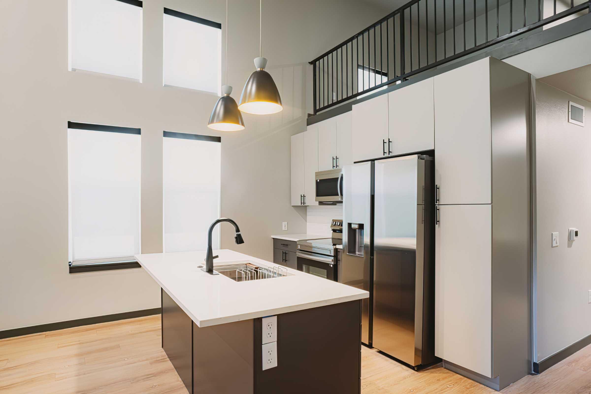 Modern kitchen featuring an island with sink, stainless steel refrigerator, stove, and microwave. The space has large windows for natural light, wood flooring, and pendant lighting hanging over the island. A loft area is visible in the background.