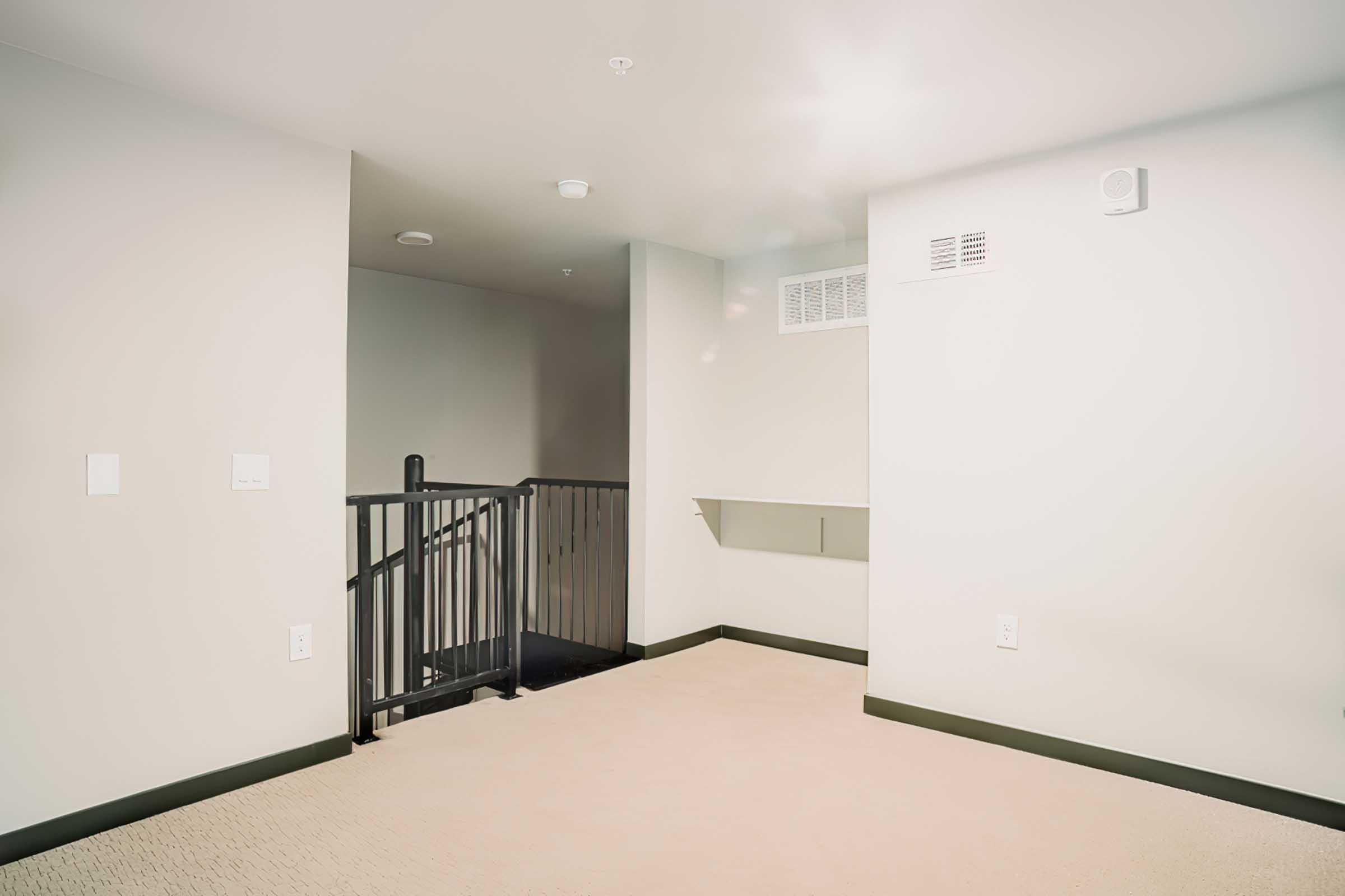 Empty room with light-colored walls and a beige carpet. A staircase is visible in the background, leading to a lower level. There is a small wall-mounted shelf on one side, and a ventilation grill on the opposite wall. The lighting is bright, creating a spacious feel.