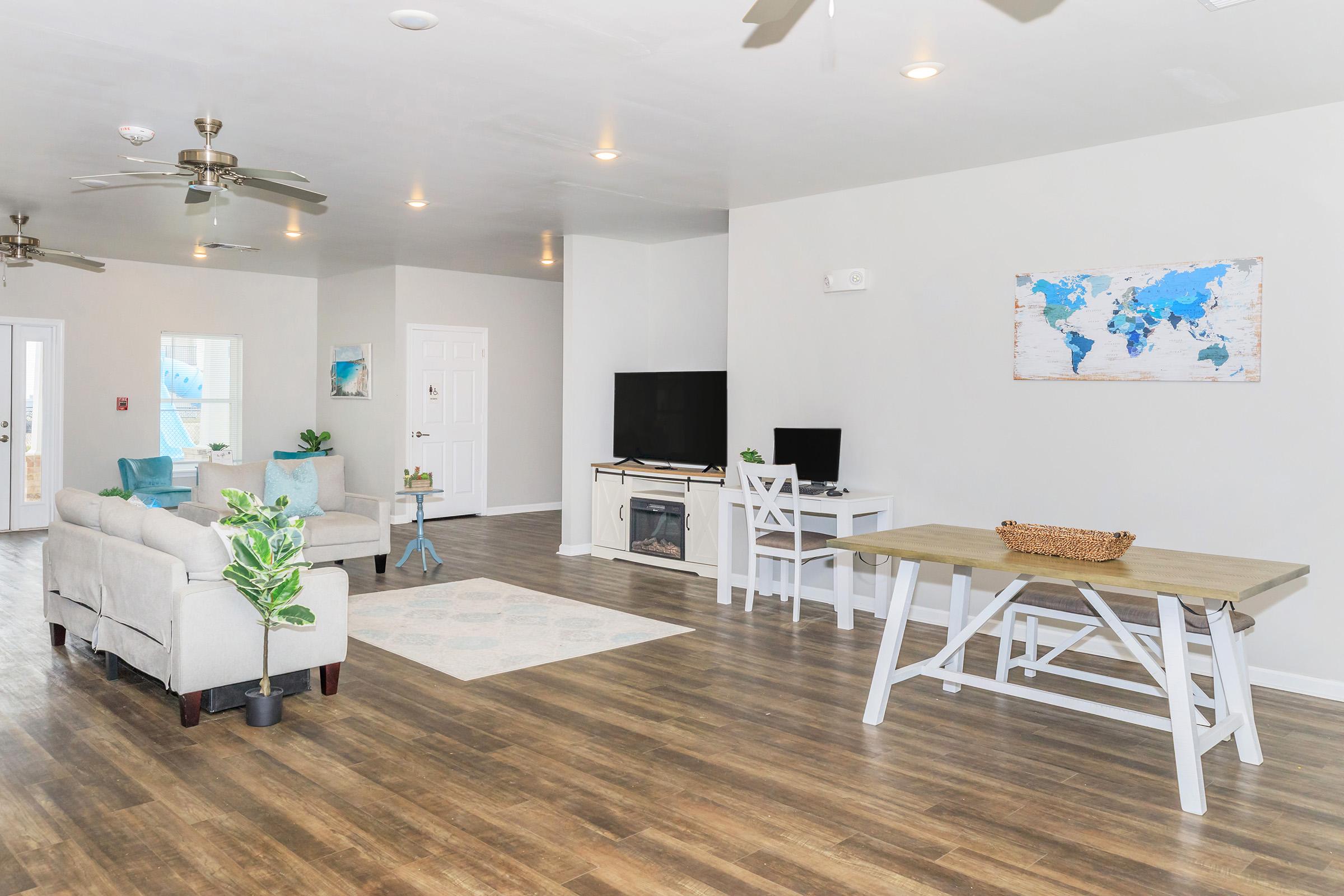 Spacious and modern living area featuring a light-colored sofa, wooden dining table, and a large TV. A world map art piece hangs on the wall, while potted plants add a touch of greenery. The room is well-lit with ceiling fans and natural light from large windows.