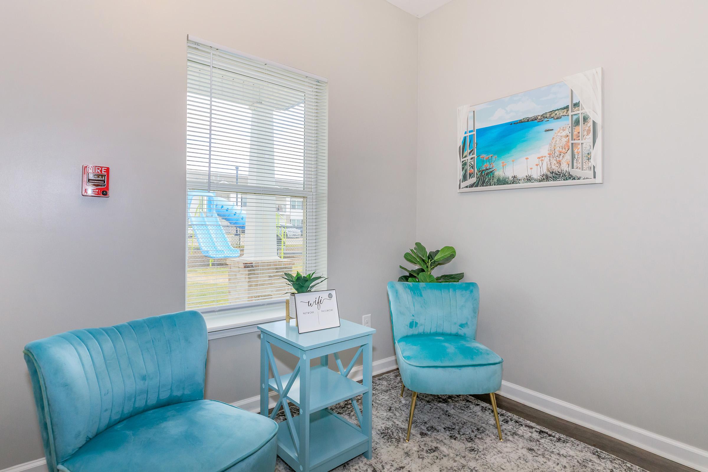A cozy corner room featuring two turquoise chairs and a small blue side table with a potted plant. A colorful painting of a beach scene hangs on the wall, and a window with blinds allows natural light to stream in. In the background, a glimpse of a playground slide is visible outside.