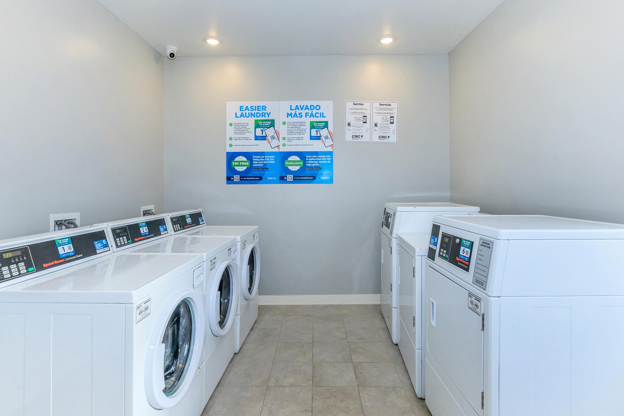 A clean and modern laundry room featuring multiple white washing machines and dryers. The walls are light gray, and there is a poster on the wall with laundry instructions in English and Spanish. Soft, bright lighting enhances the space, creating an inviting atmosphere for laundry tasks.