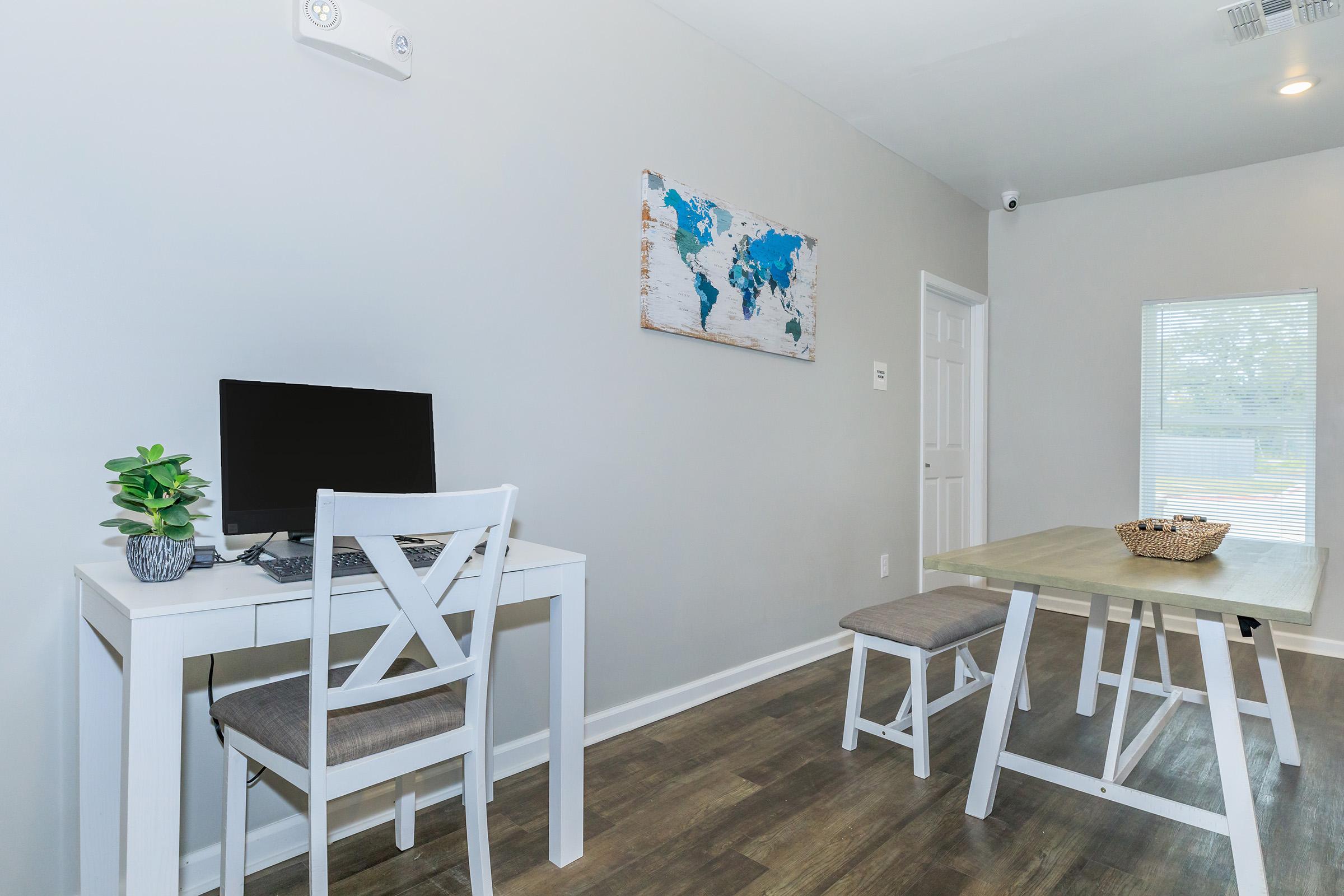 A modern home office setup featuring a white desk with a computer, a green plant, and a light-colored chair. Adjacent is a dining area with a wooden table and a bench. A world map hangs on the wall, and a window provides natural light, enhancing the bright and airy atmosphere.