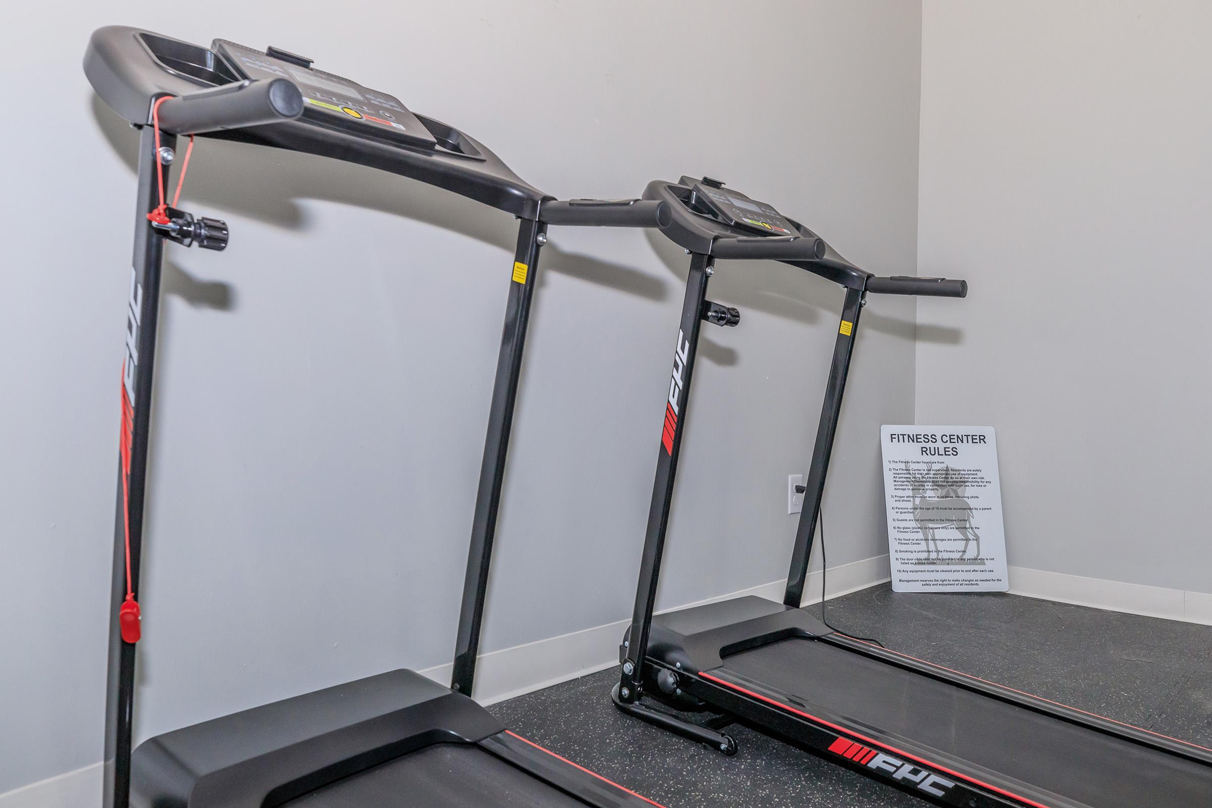 Two black treadmills are positioned side by side against a gray wall. A sign detailing fitness center rules is posted nearby, emphasizing safety and usage guidelines. The floor is a dark, textured surface, and the treadmills have digital displays and buttons for various settings.