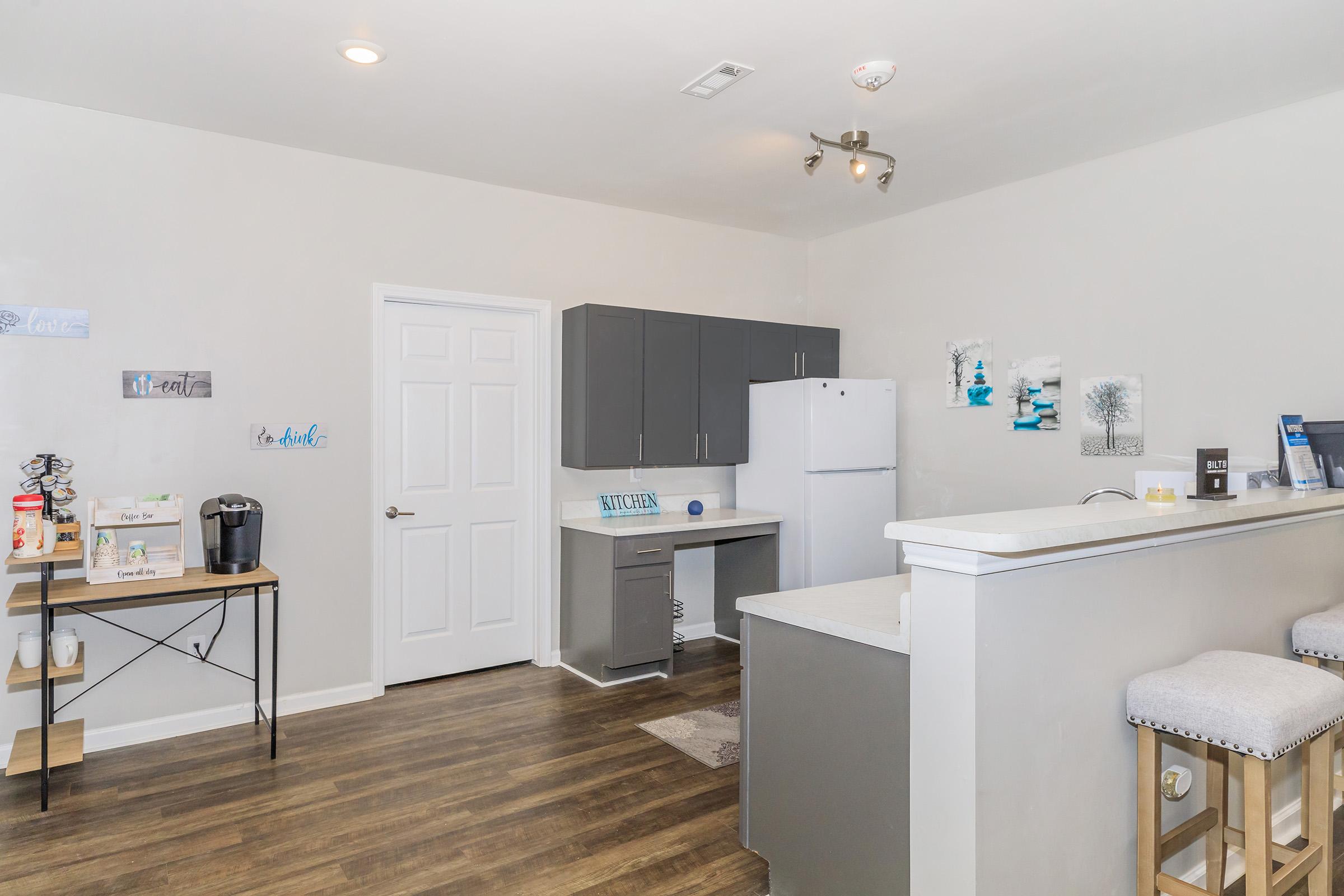 A modern kitchen and living area featuring gray cabinetry, a white refrigerator, and a coffee machine on a small table. The space has a neutral color palette with wooden flooring, and there are decorative elements on the walls, including artwork and inspirational quotes. Barstools are positioned near the kitchen counter.