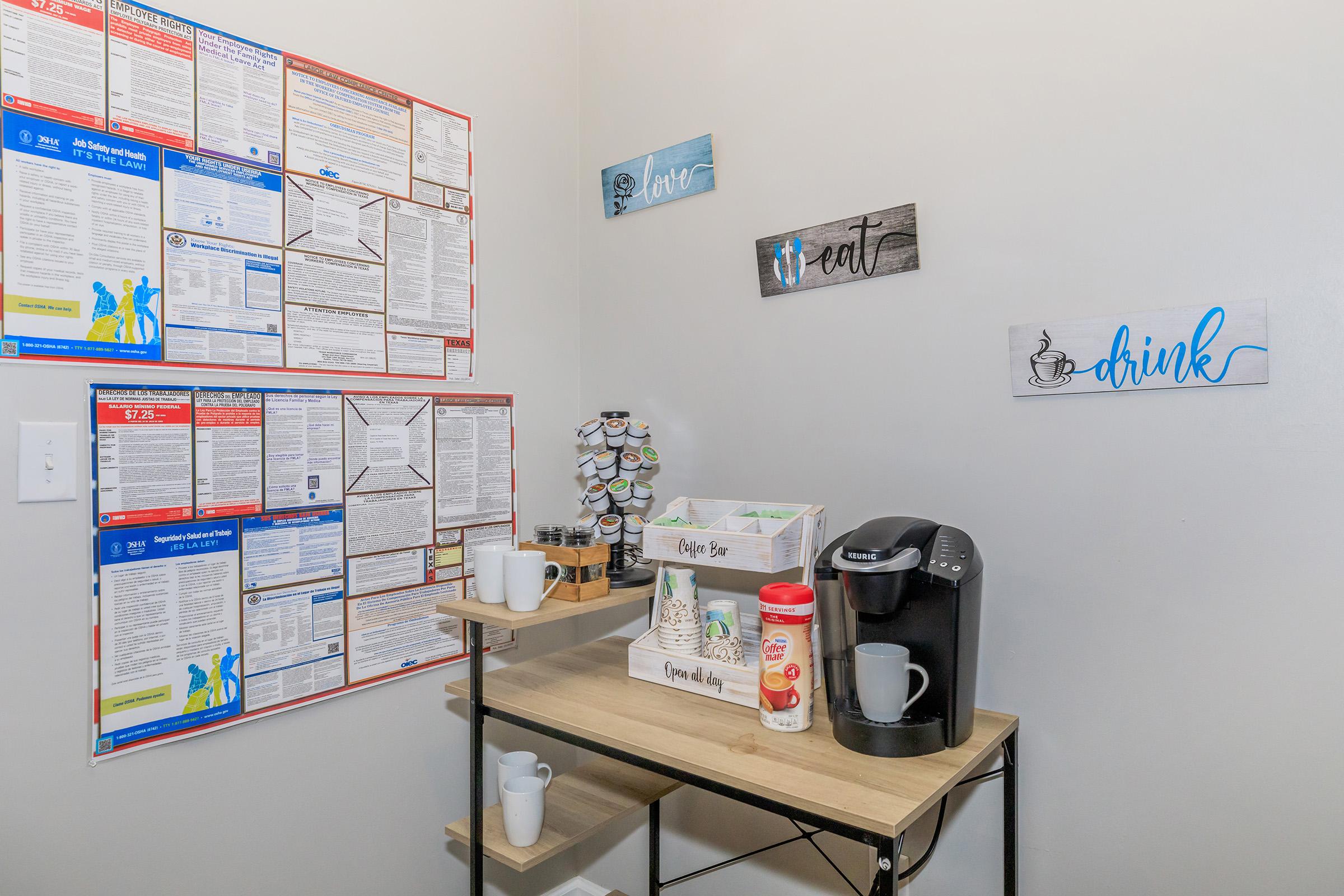 A small office break room featuring a coffee station with a Keurig machine, cups, and sugar. The wall displays various informational posters. Decorative signs on the wall encourage relaxation with the words "love," "eat," and "drink." A spinning rack holds coffee pods nearby.