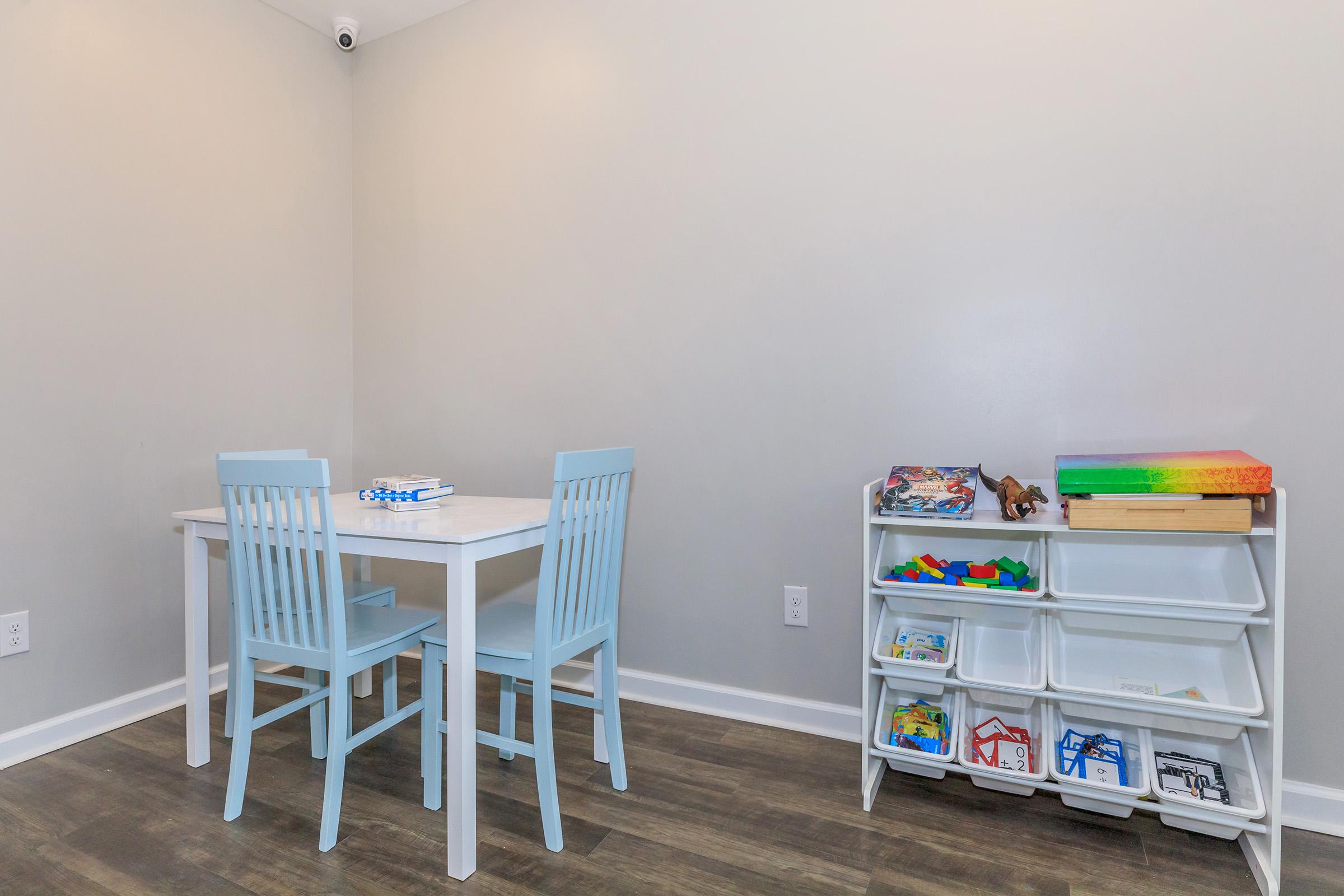 A light and simple playroom with a small white table and two blue chairs. There is a shelf with colorful storage bins containing toys and games. The walls are a neutral color, and a security camera is visible in the ceiling corner. The floor features wood-like laminate.