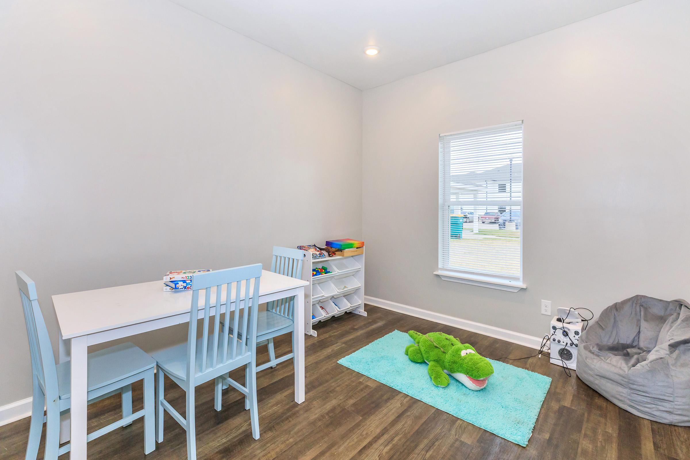 A bright, child-friendly room featuring a light blue table with four matching chairs, a white storage shelf filled with toys, a green plush alligator on a blue rug, and a comfortable bean bag chair. Natural light streams in through a window, illuminating the playful and inviting space.
