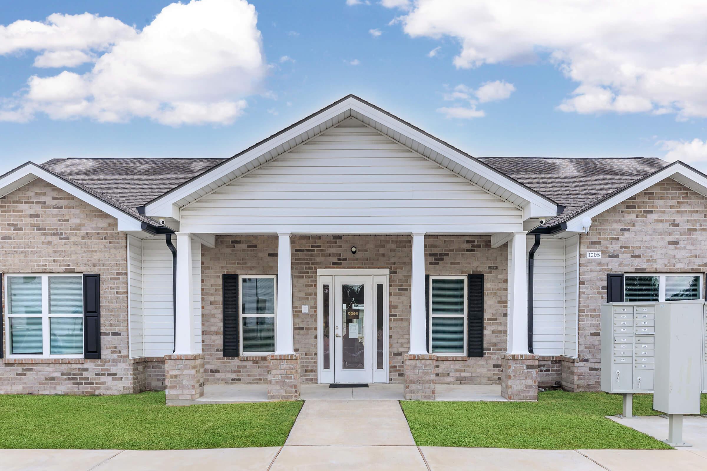A modern single-story brick house with a white exterior and a gable roof. It features a covered porch with white columns, large windows, and a front door. A row of mailboxes is visible on the right side, and green grass lines the walkway leading to the entrance. The sky is clear with a few clouds.