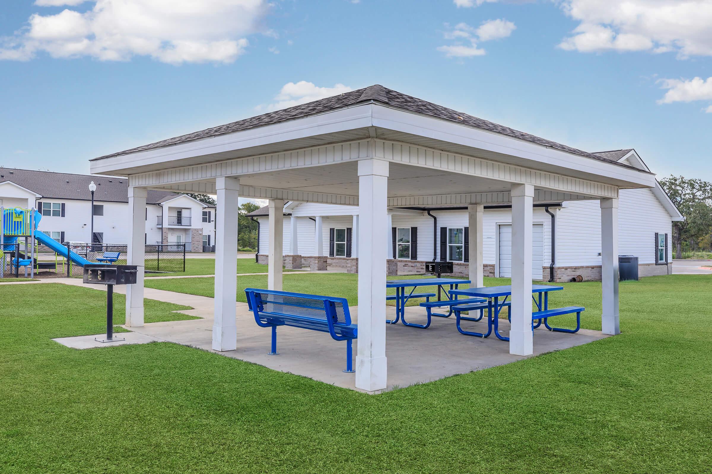 A covered picnic area featuring blue tables and benches, situated on a grassy lawn. In the background, there are residential buildings and a playground with slides and climbing structures, set against a clear blue sky with fluffy clouds.