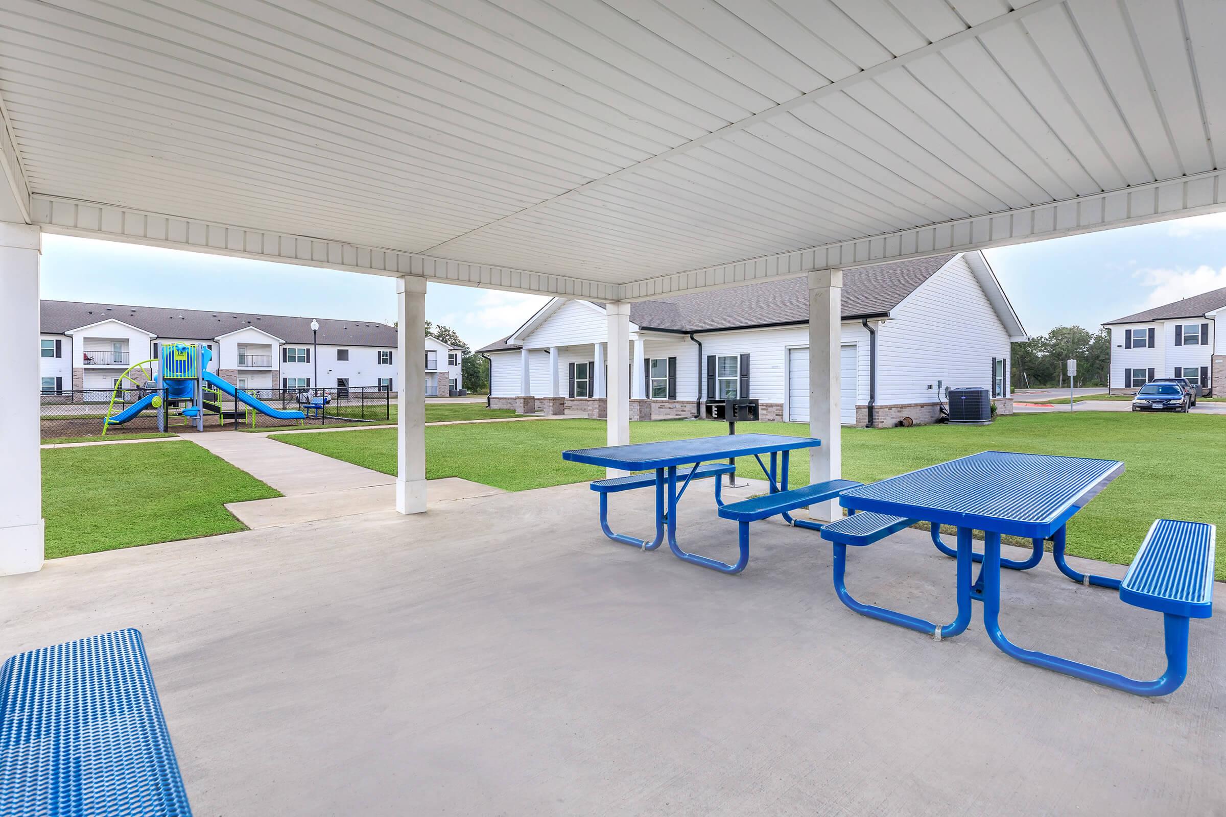 A covered picnic area with blue tables and benches, overlooking a grassy space and a playground with a slide and climbing structure. Nearby, there are residential buildings with white exteriors. The sky is overcast, indicating a cloudy day.
