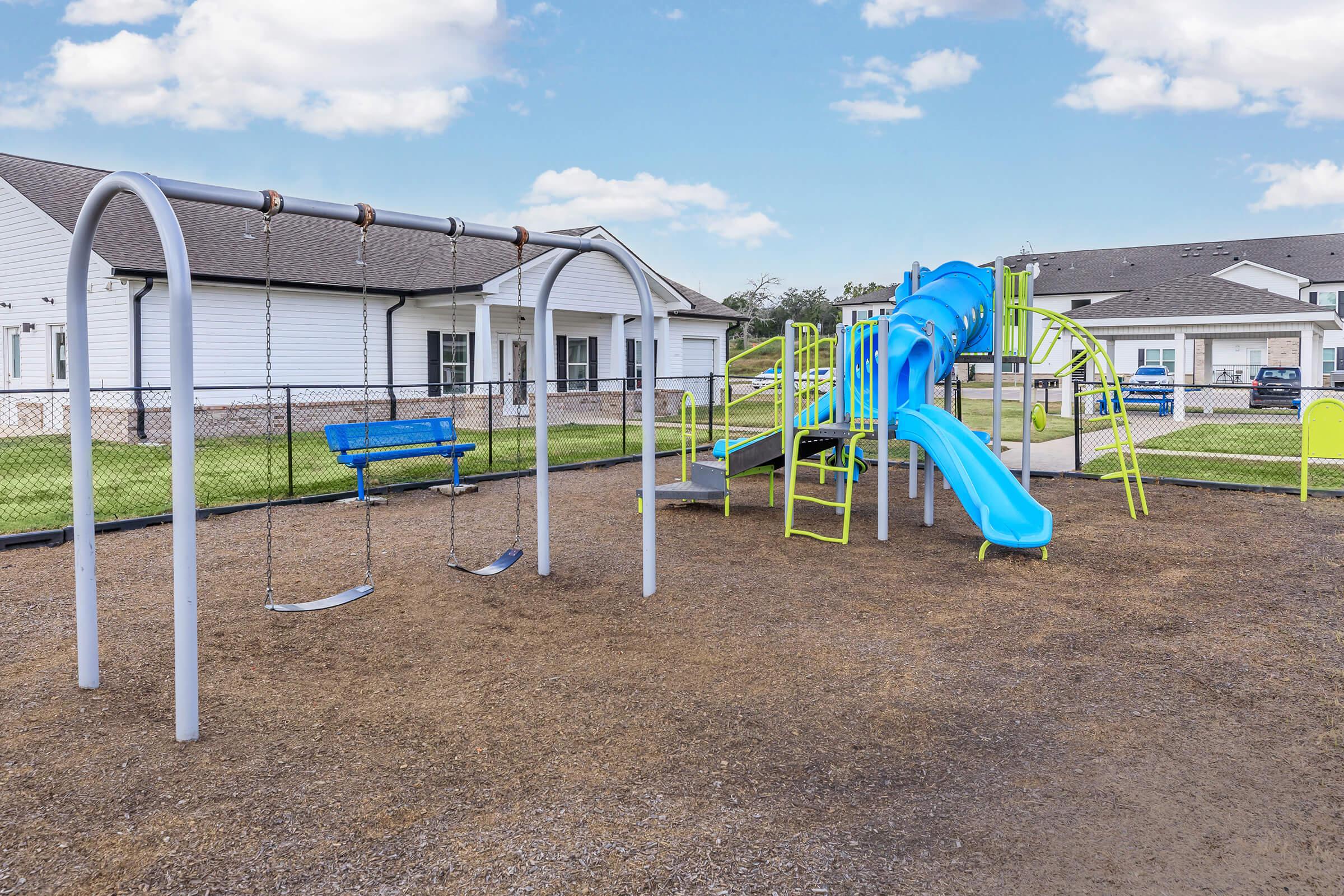 A colorful playground featuring a blue slide, green climbing structure, swings, and a fenced area. The background includes white buildings and a grassy lawn under a blue sky with scattered clouds.