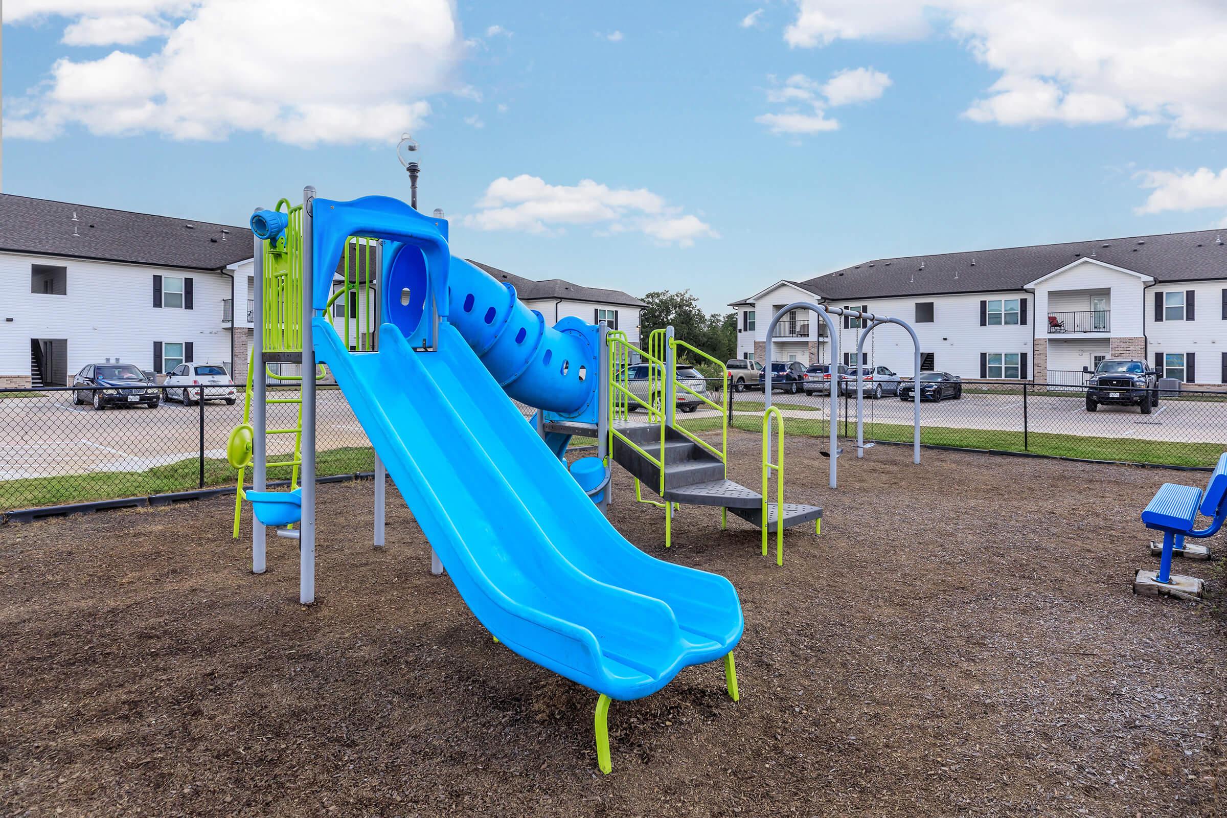 Colorful playground equipment featuring two blue slides and a climbing structure, set on a gravel surface. In the background, there are several apartment buildings and a parking area visible under a clear blue sky. The playground is enclosed by a fence.