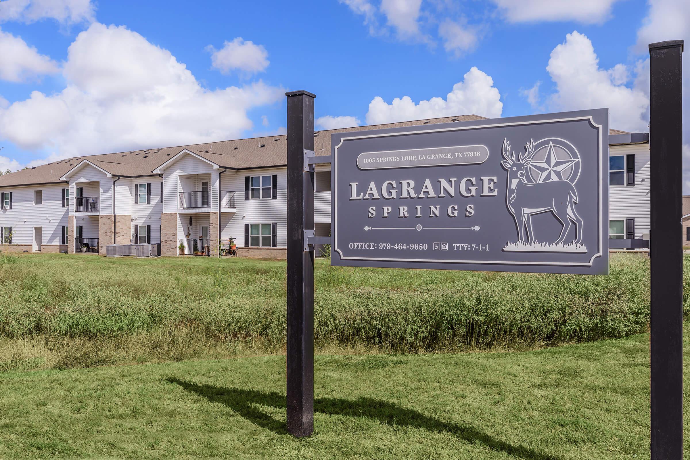 Sign for LaGrange Springs, featuring a deer silhouette. The sign includes contact information and is placed in front of a multi-story residential building surrounded by green grass under a blue sky with clouds.