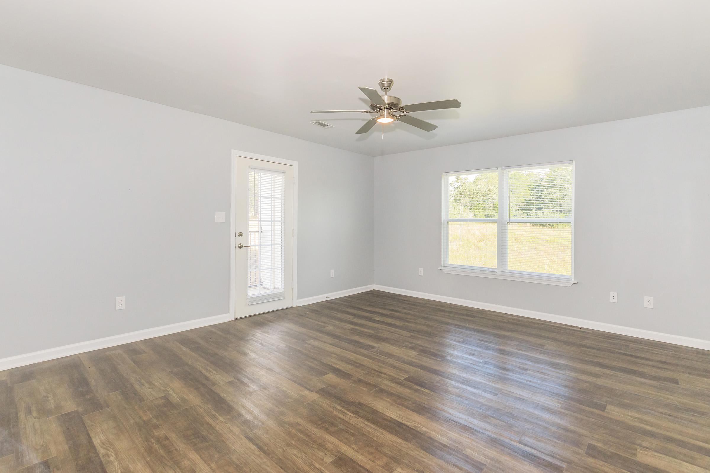An empty room with light gray walls and a large window on one side. The floor features wood-like laminate, adding warmth to the space. A ceiling fan is installed, and a door leads outside. Natural light brightens the interior, emphasizing the room's spaciousness and potential for various uses.