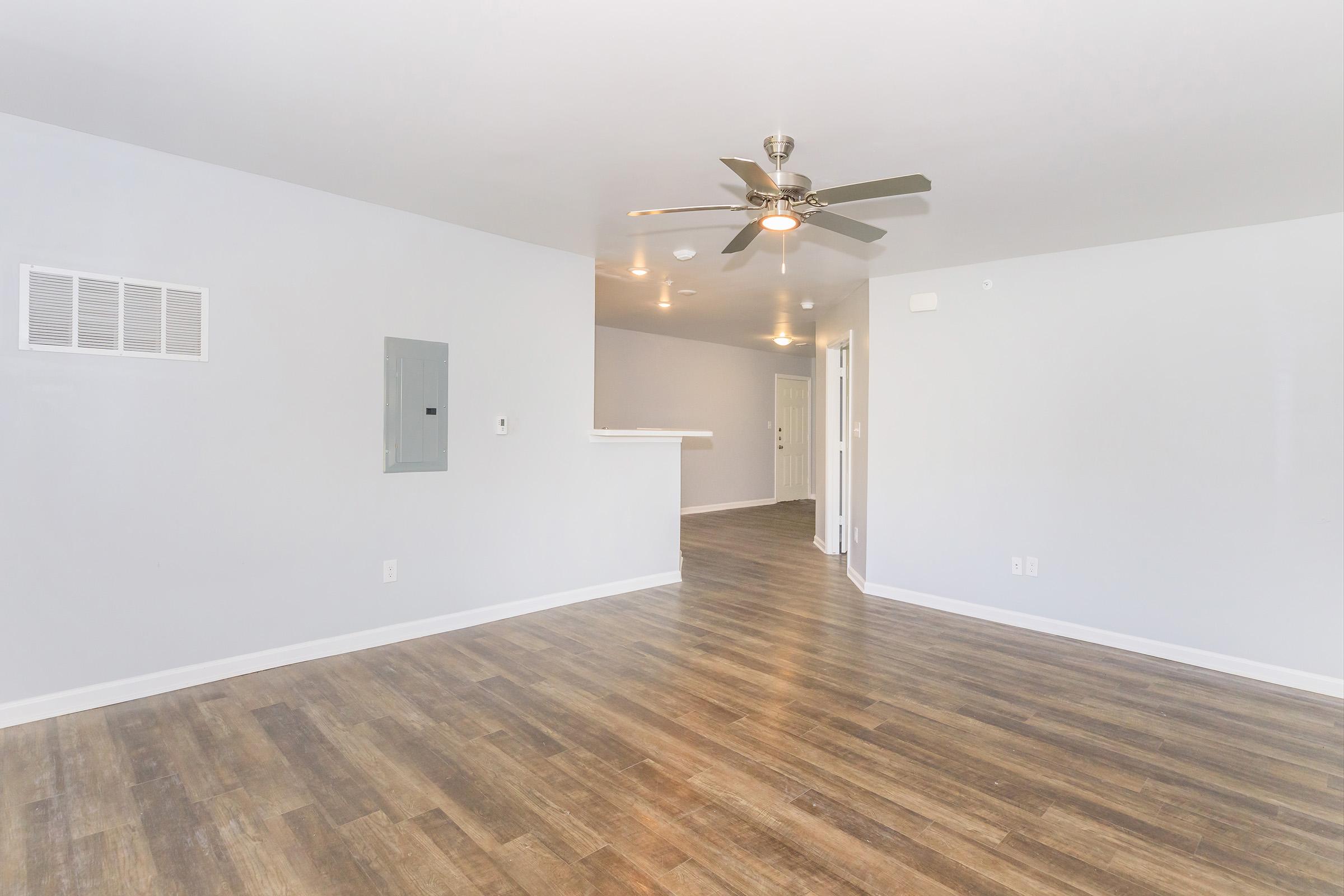 Modern, spacious living area featuring light gray walls, a ceiling fan, and wood-like flooring. An electrical panel is mounted on the wall, with a door leading to another room in the background. The space is well-lit with recessed lighting, creating an inviting atmosphere.