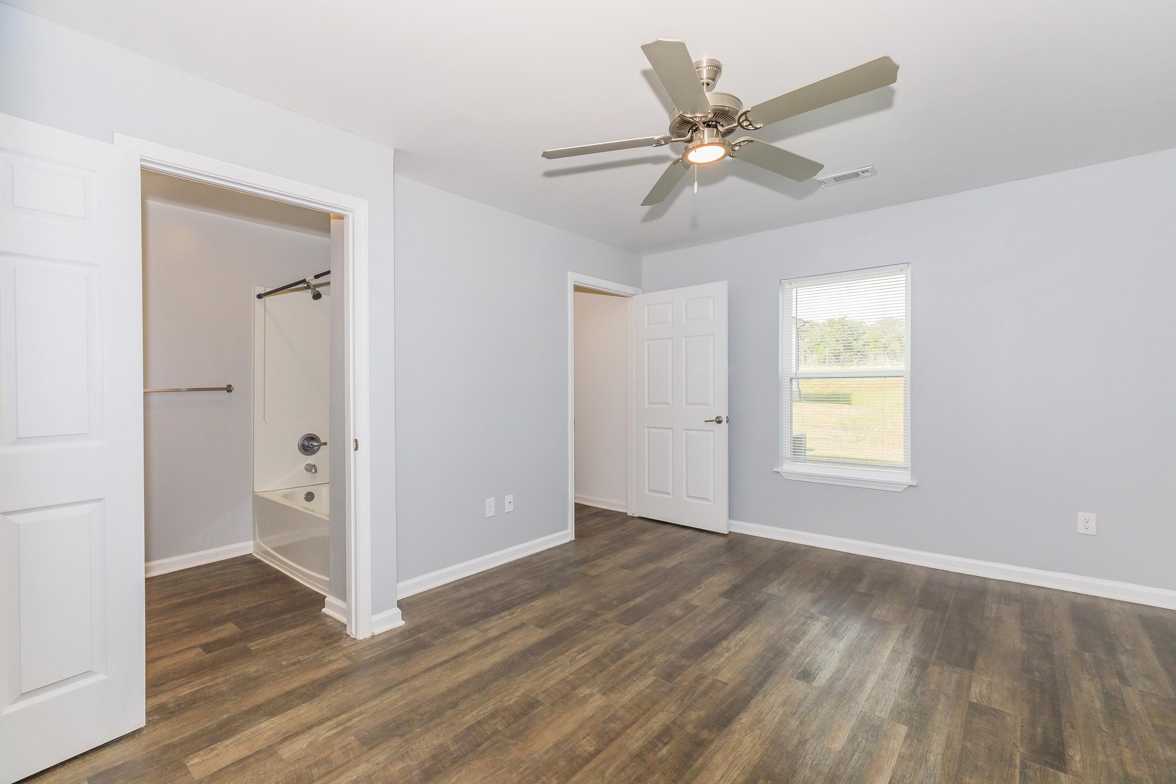 A bright room featuring light gray walls and a ceiling fan. A door opens to a closet area with a shower stall visible. There is a window providing natural light, and the floor is covered in wooden planks. Two doors are visible, leading to different spaces.