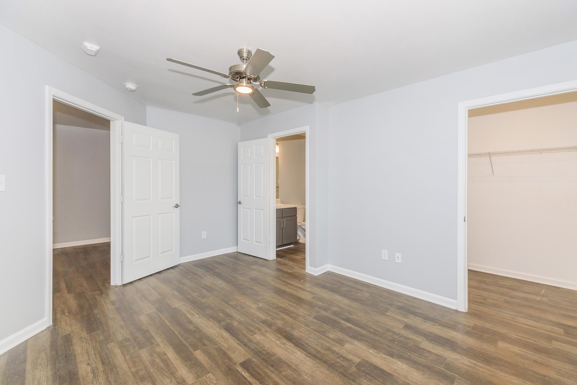 A clean, modern room with light gray walls and wooden flooring. It features a ceiling fan and two open doors leading to adjacent rooms. One door leads to a bathroom area, while the other opens into a closet space, providing a spacious and neutral environment suitable for various decor styles.