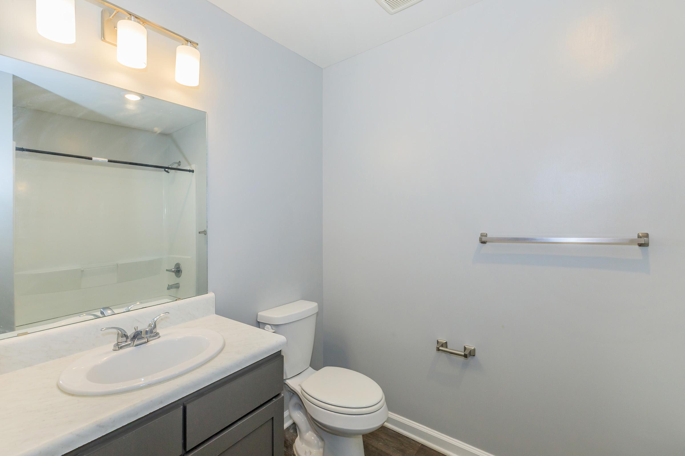 A well-lit bathroom featuring a modern sink with a marble countertop, a toilet, a mirrored wall, and a towel bar. The walls are painted light gray, and there is a glass shower enclosure visible in the background. The overall design is clean and minimalist.