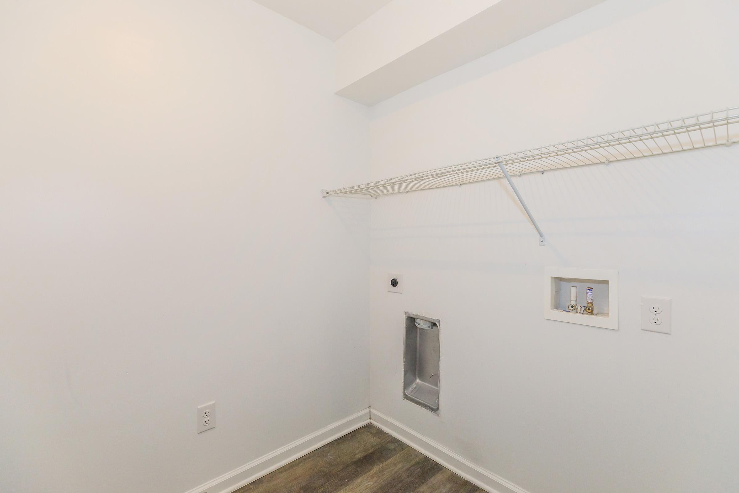 Empty room with white walls and a light grey floor. There is a wire shelf mounted on the wall, and a small access panel in the lower right corner. The space is well-lit and free of furniture or decor, creating a minimalist appearance.