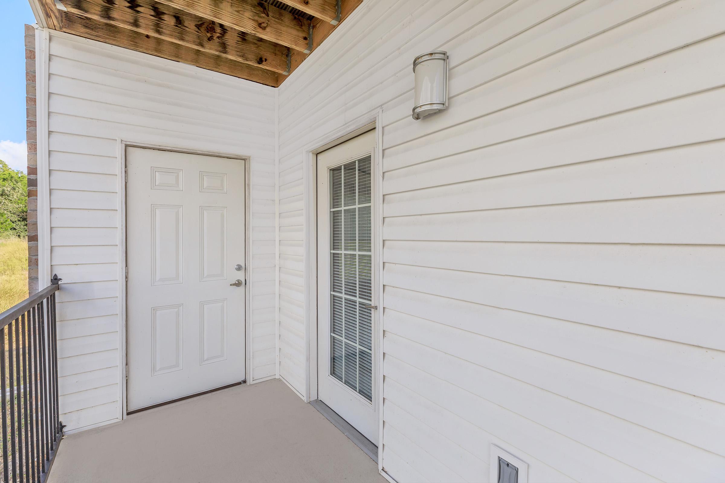 A white exterior of a building featuring a main entrance door and a sliding glass door. The walls have horizontal siding, and a covered porch with wooden beams is visible above. A light fixture is mounted near the main door, and there are grassy fields in the background.