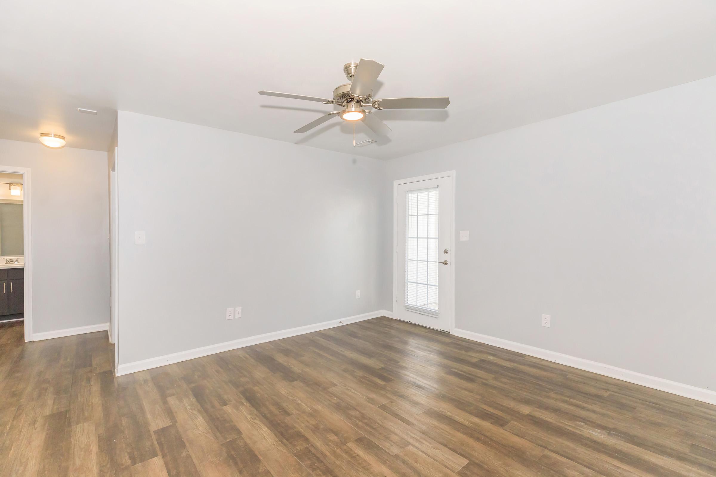A bright and empty room with light gray walls and a ceiling fan. The floor features brown laminate or hardwood, creating a warm look. A door leads to an exterior space, with natural light coming through a nearby window. The room has a modern, minimalist aesthetic.