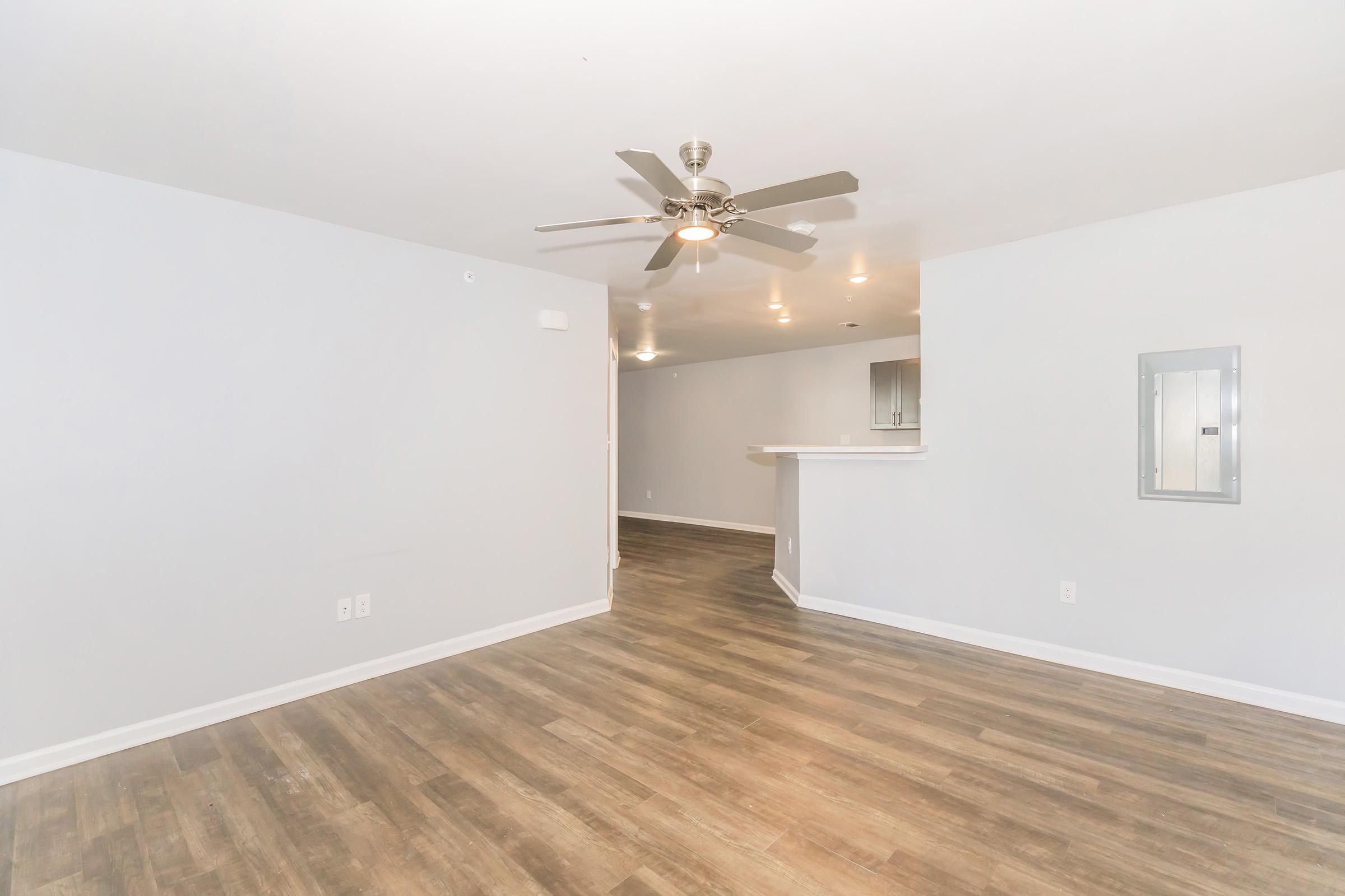 A spacious, modern living area featuring light gray walls and a ceiling fan. The floor is made of light wood, and there's a bar counter leading to a kitchen area in the background. The space is well-lit with recessed lighting, creating a bright and inviting atmosphere.