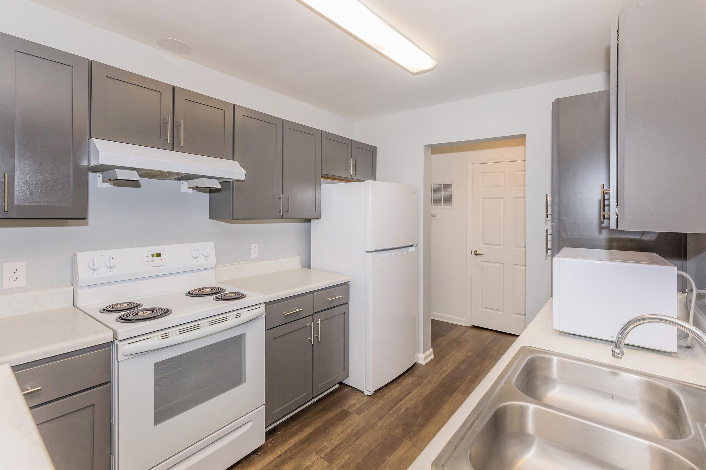 Modern kitchen featuring grey cabinets, white countertops, a white refrigerator, and a white stove with an oven. The room is illuminated by overhead lighting, and there is a double sink and a door leading to another space. The flooring is a dark wood laminate.