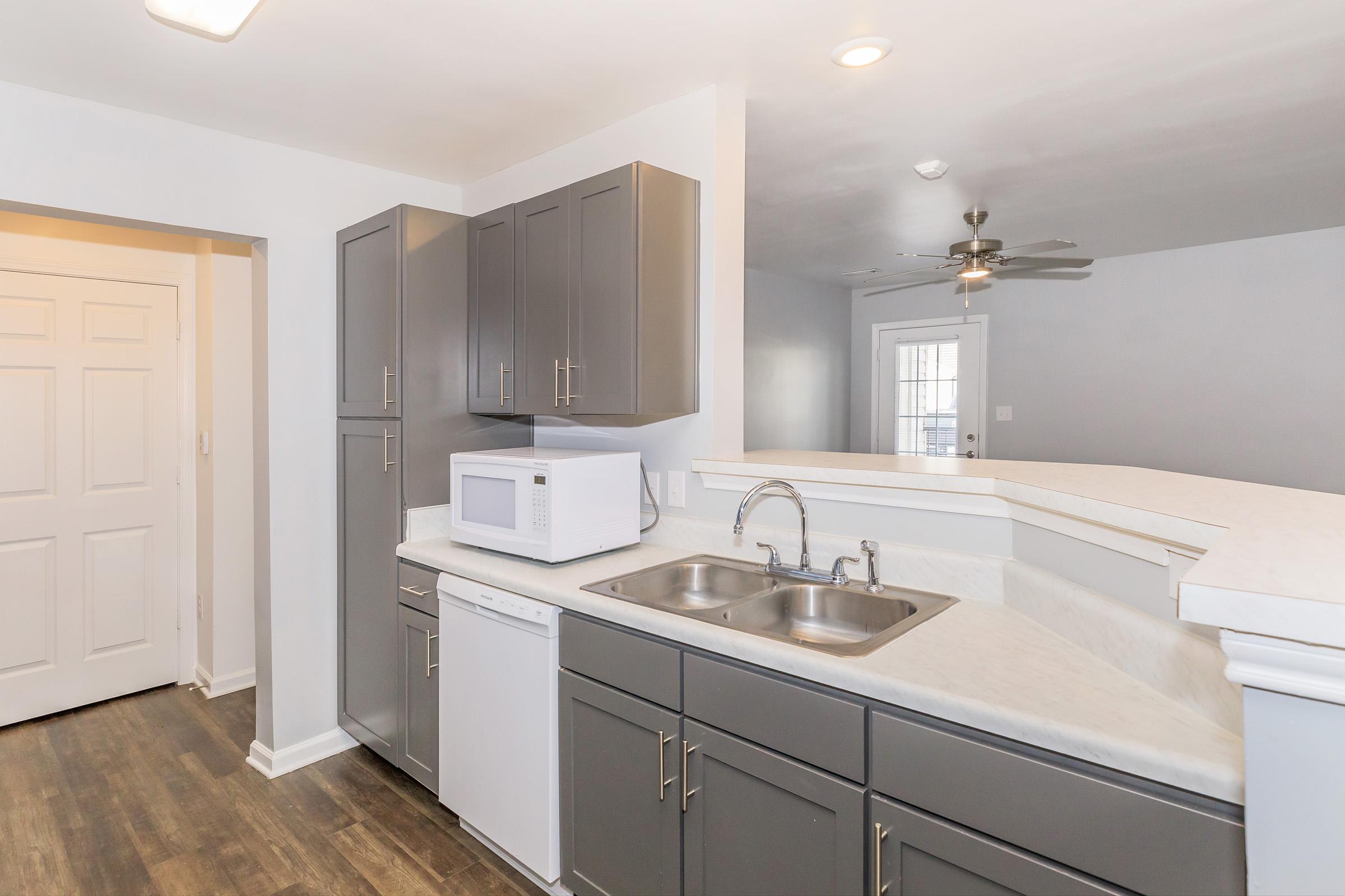 A modern kitchen featuring dark gray cabinets, a double sink, a microwave, and a dishwasher. The countertops are light-colored, and the space includes a doorway leading to another room. Natural light enters through a nearby window, complementing the contemporary design.