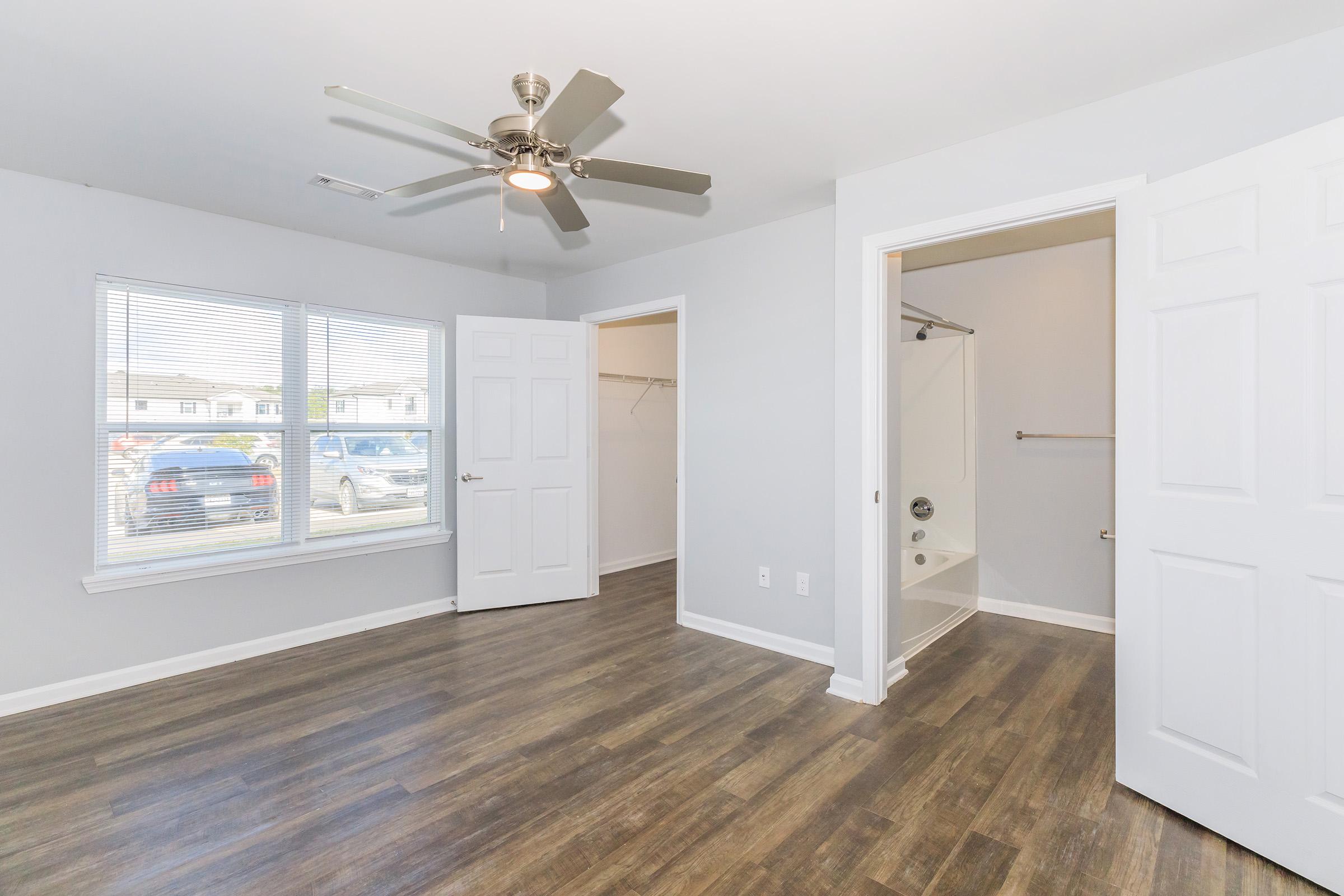 Spacious room featuring a ceiling fan, light gray walls, and a large window with blinds. There's a closet area to the left and an open bathroom space with a shower on the right. The floor is a modern wood-style laminate, creating a warm and inviting atmosphere.