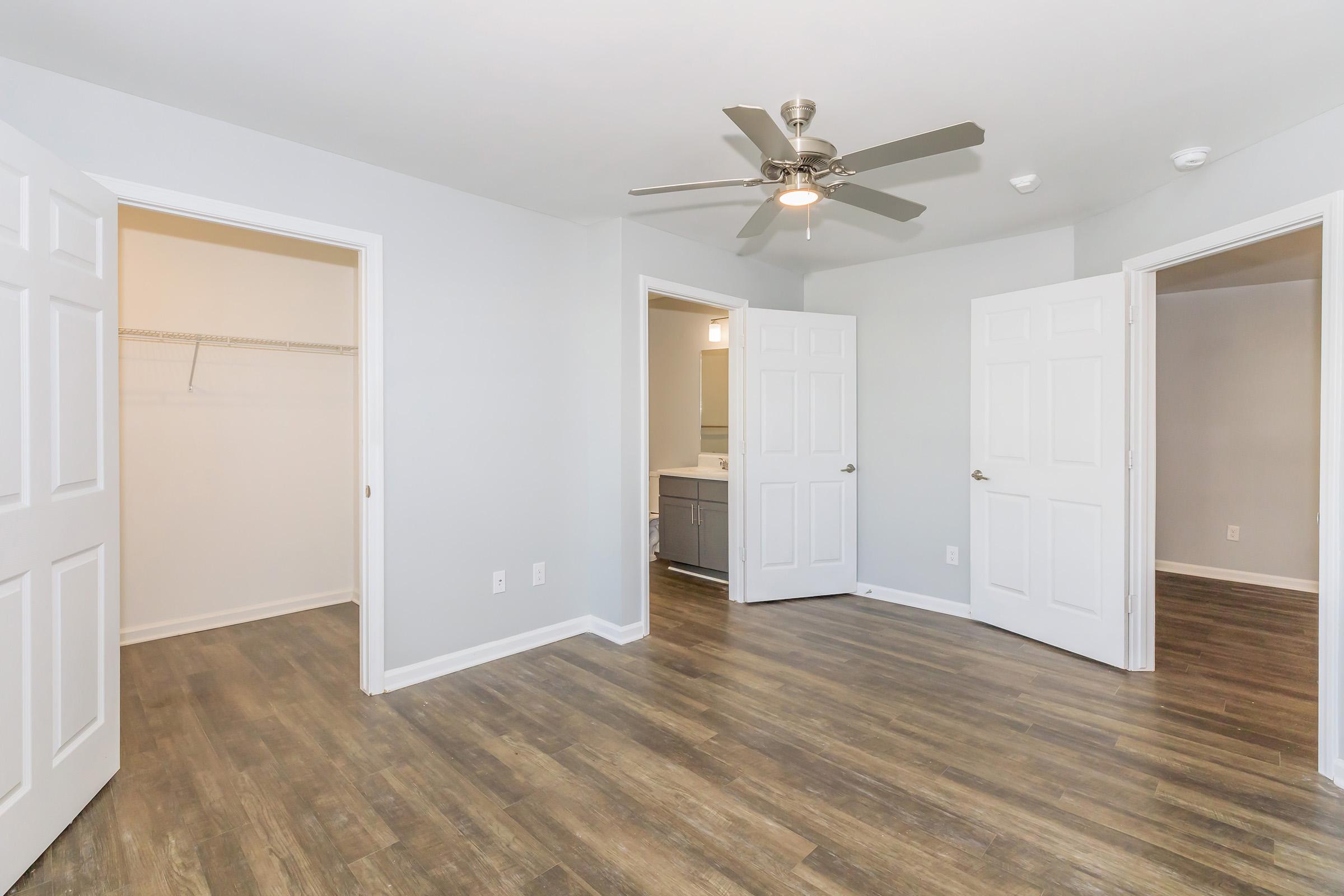 A spacious room featuring light gray walls, a ceiling fan, and hardwood-style flooring. There are three doors—two leading to closets and one to a bathroom area. The layout is open and airy, with ample natural light.