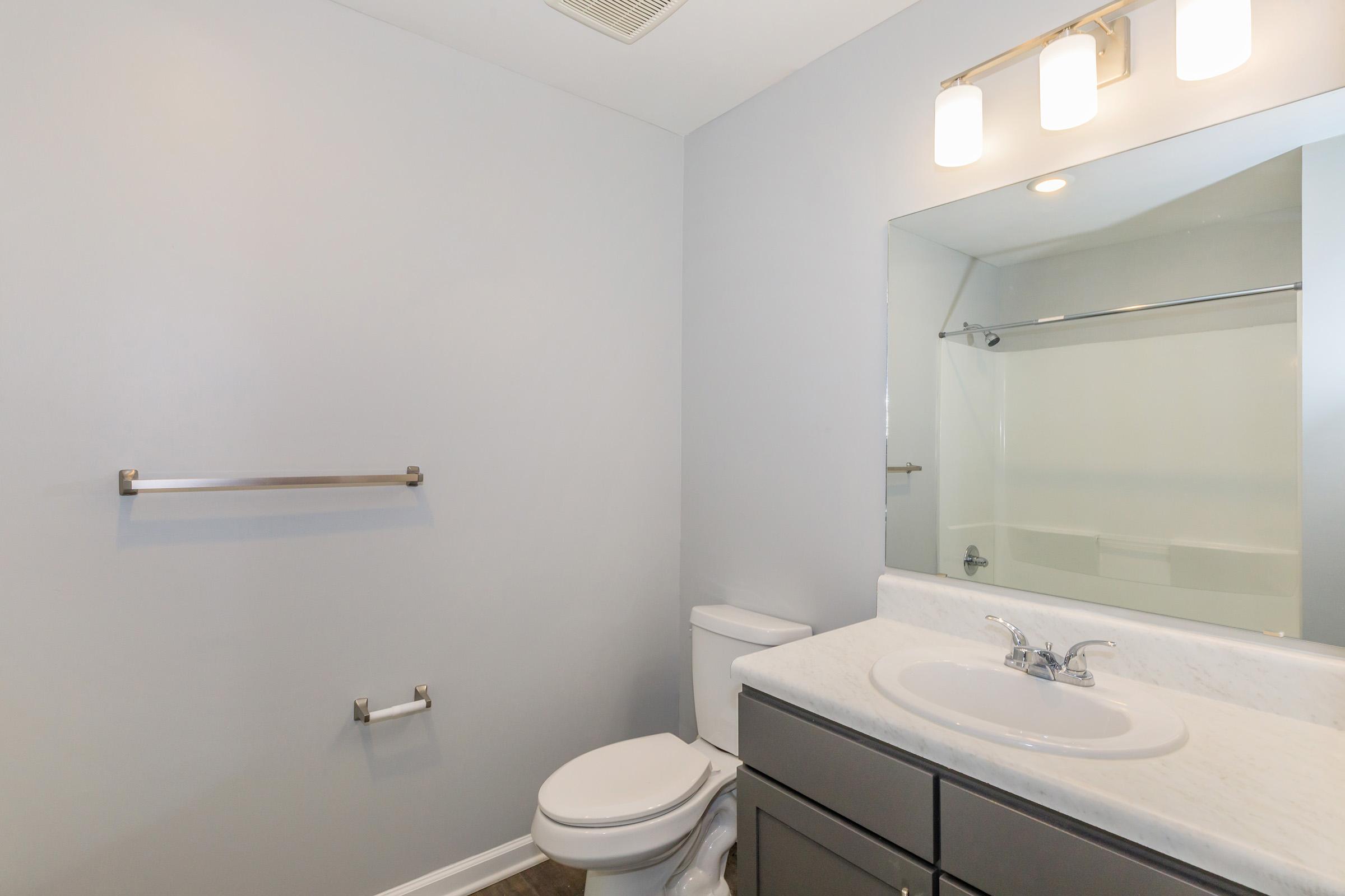A modern bathroom featuring a white toilet, a sleek vanity with a round sink, and a large mirror above it. The walls are painted in a soft gray color, and there is a metal towel rack on the wall. The flooring is dark, contrasting with the lighter elements of the space.