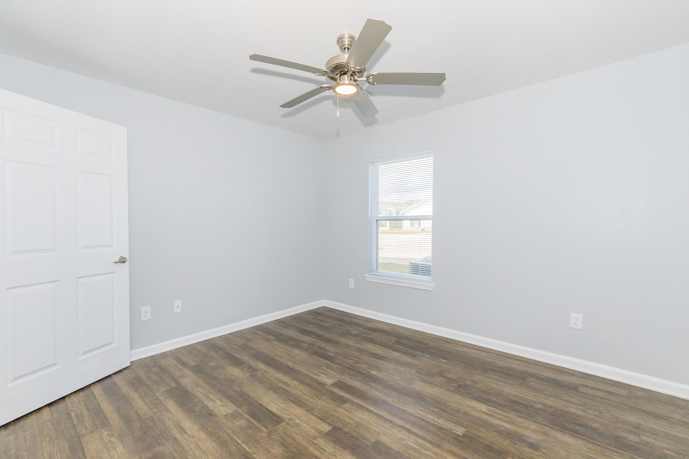 Empty room featuring light gray walls, a ceiling fan, and a window with blinds. The flooring is wood-like and there is a closed white door on the left side. The overall atmosphere is bright and spacious, ideal for customization.