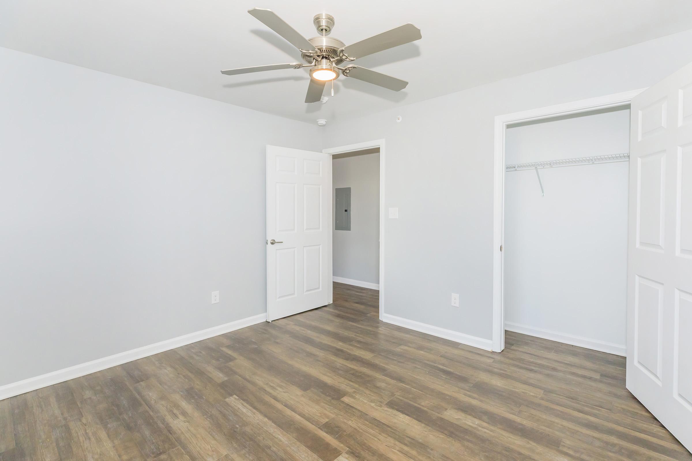 A well-lit, minimalistic room with gray walls and a ceiling fan. The room features two doors—one leading to a closet—and a wood-like floor. The space is empty, creating a clean and spacious atmosphere, ideal for various uses.