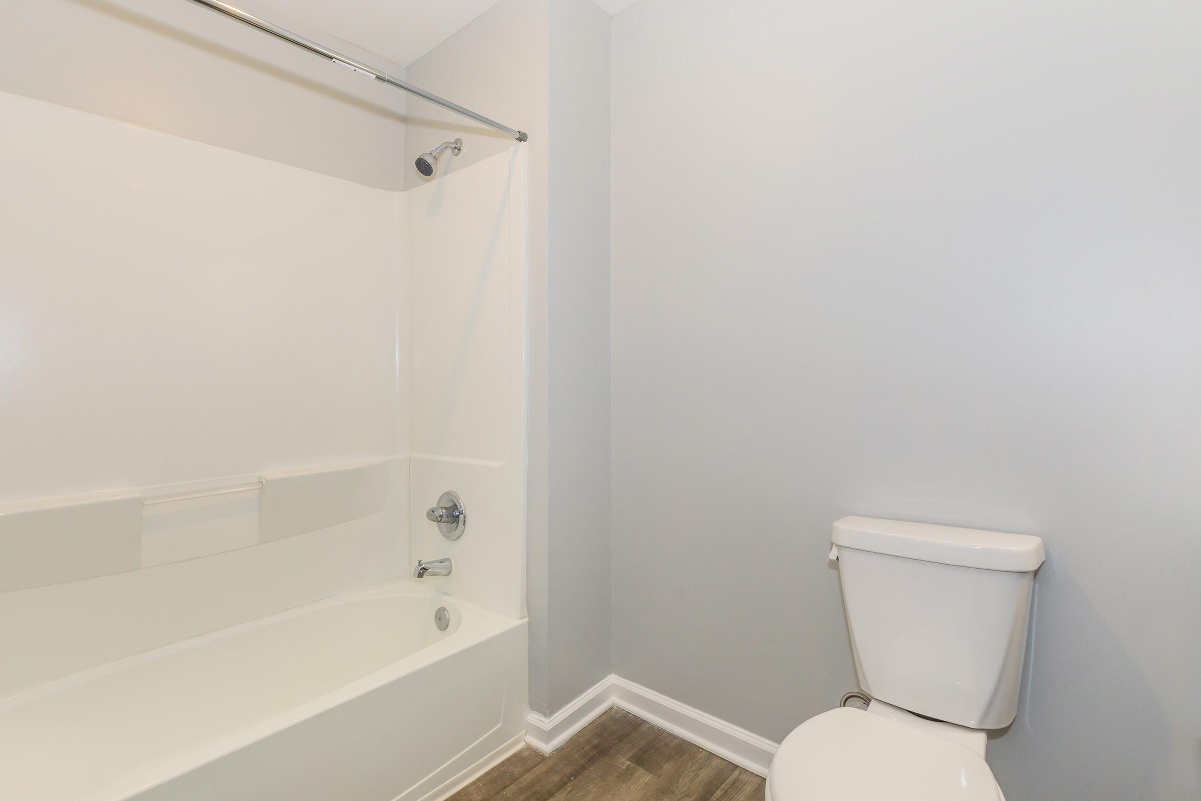 A clean and simple bathroom featuring a white bathtub with a shower curtain rod, a chrome shower faucet, and a white toilet. The walls are painted in light gray, and the flooring is a light wood finish, creating a minimalist and modern aesthetic.