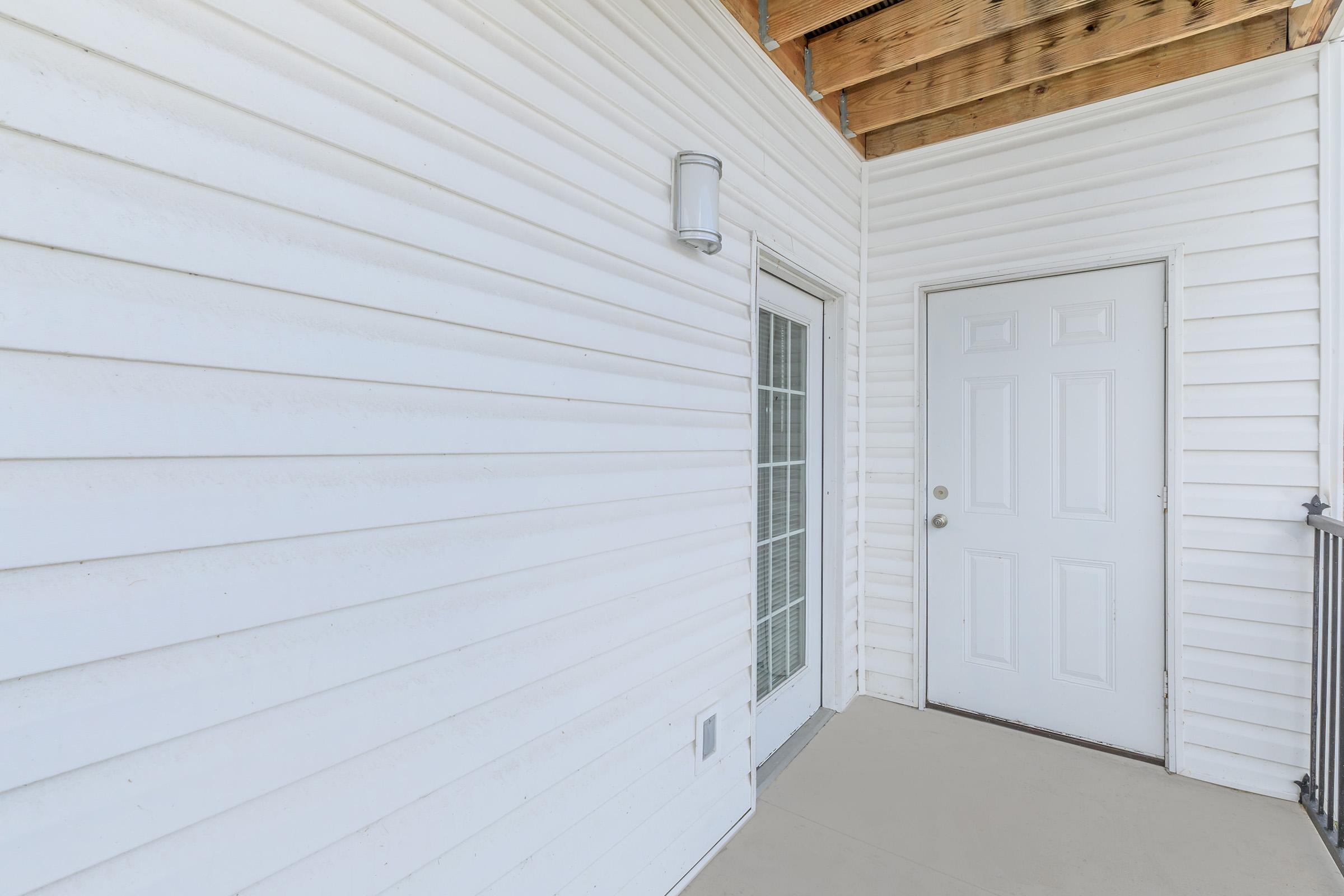 Exterior view of a porch featuring white siding, a door with a window, and a light fixture on the wall. The space is well-lit and has a simple, clean design. The floor is light-colored, and the ceiling showcases wooden beams.