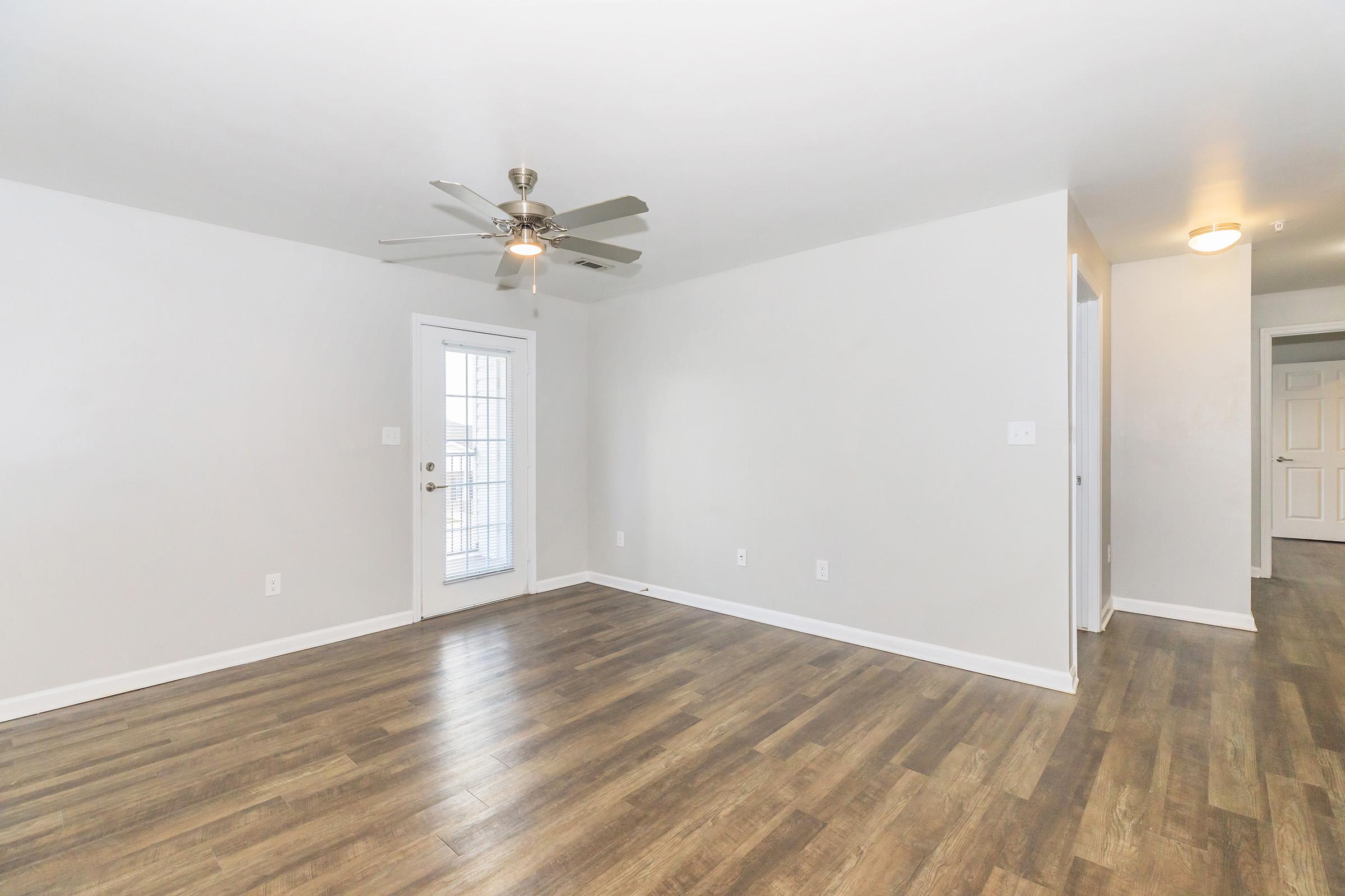 A spacious room with light-colored walls and wood-style flooring. It features a ceiling fan and a door leading outside. There are no furnishings, creating an open and clean aesthetic. A hallway is visible on the right, leading to additional spaces.
