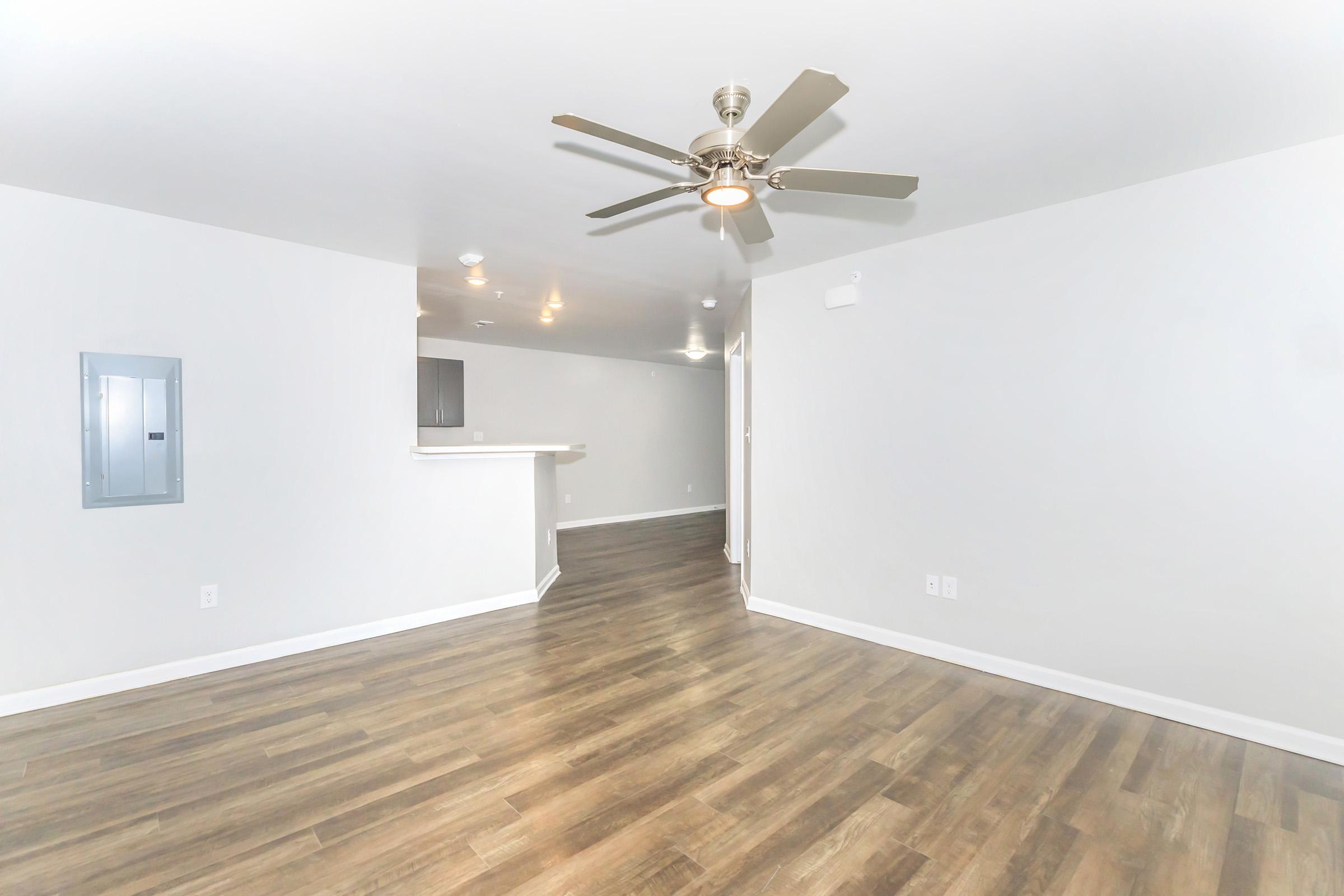 An empty room with light-colored walls and a wooden floor. A ceiling fan is mounted in the center, and a doorway leads to another area. A small electrical panel is visible on the left wall. The space appears bright and clean, suggesting it could be a living room or common area in an apartment.