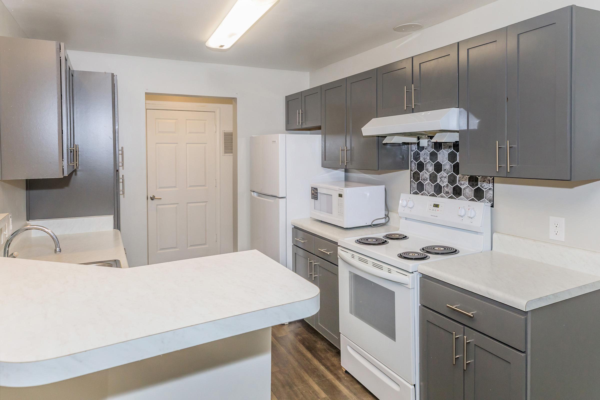 A modern kitchen featuring gray cabinetry, a white stove, and microwave, along with a refrigerator. The countertop is light-colored, and there's a decorative tile backsplash behind the stove. The space is well-lit with overhead lighting and has a doorway leading to another room.