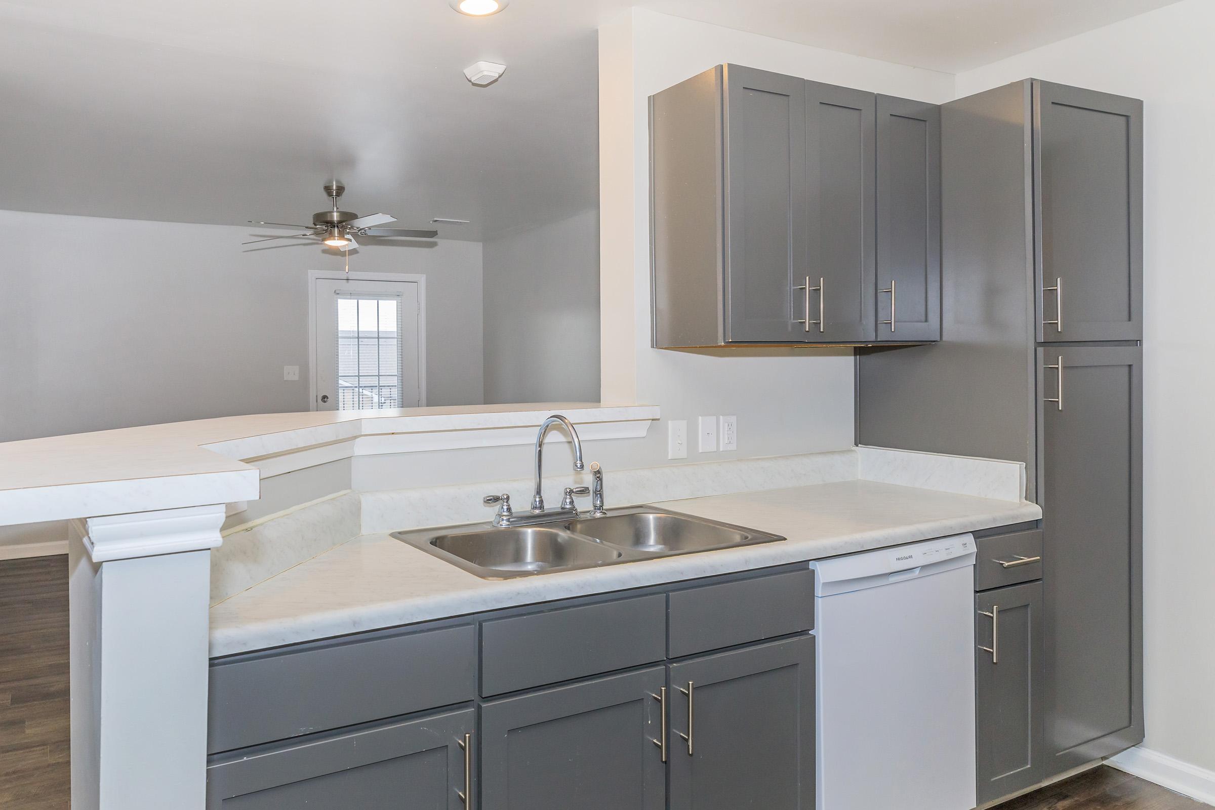 Modern kitchen featuring grey cabinets, a double sink, and a dishwasher. The countertop is light-colored, and there is a ceiling fan visible in the background. The open space leads to a living area with natural light entering through a doorway.